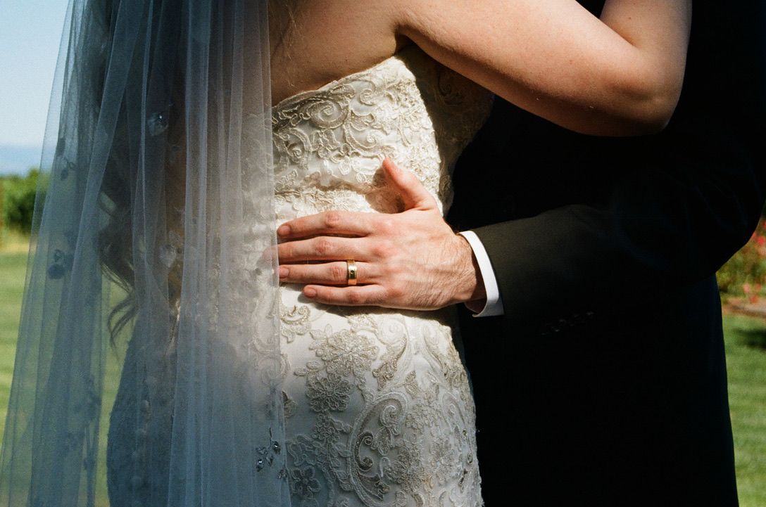 Groom's hand with wedding ring embracing bride in lace wedding dress and veil during outdoor ceremony.