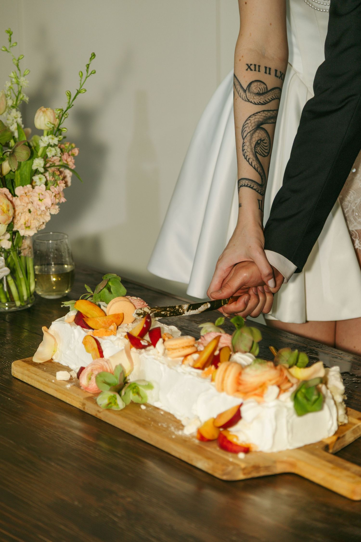 Couple with tattoos cutting a white cream cake topped with fresh peaches and fruit on a wooden board at a wedding.