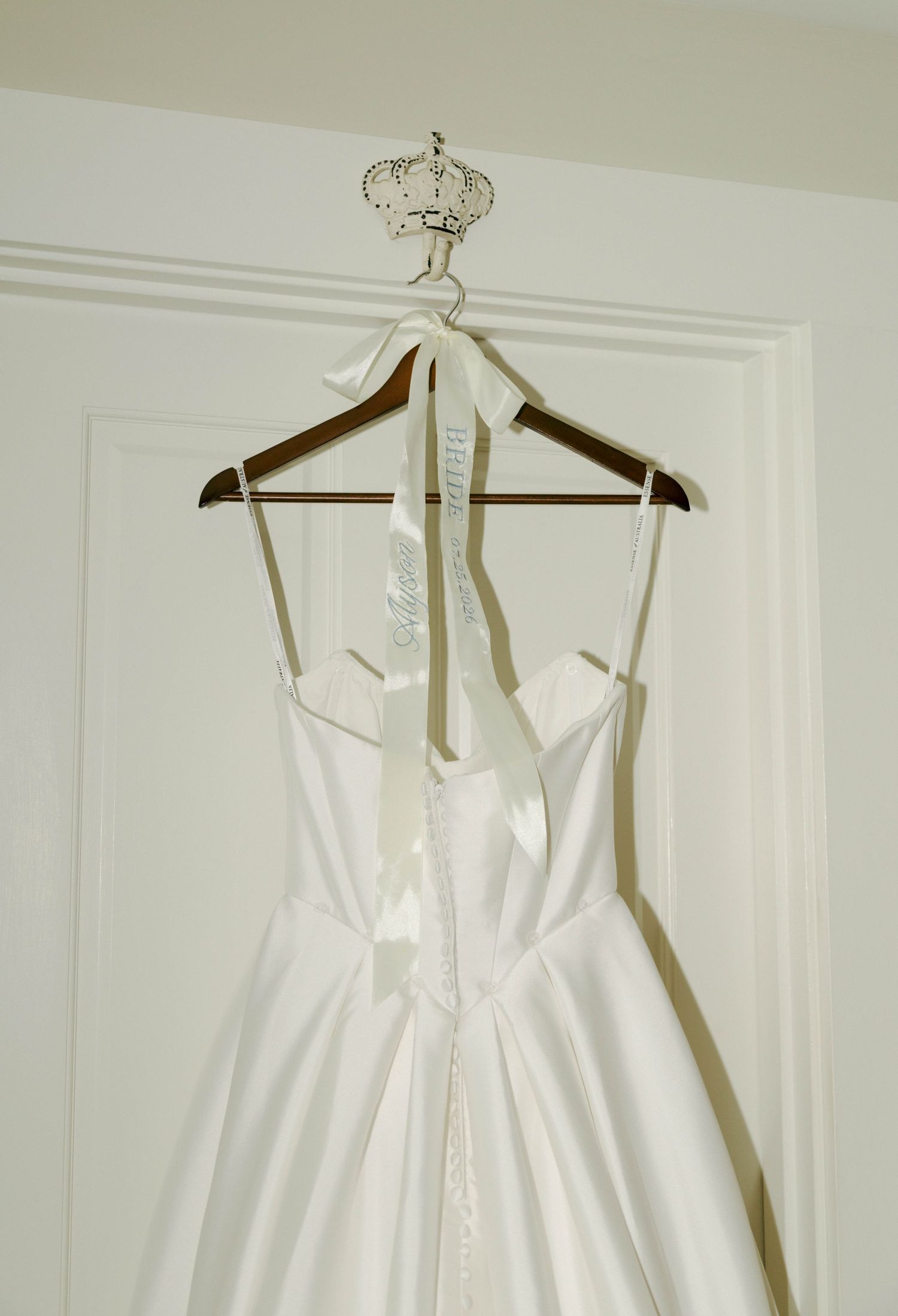Elegant white satin wedding dress hanging on a wooden hanger with ribbon details against a white door.