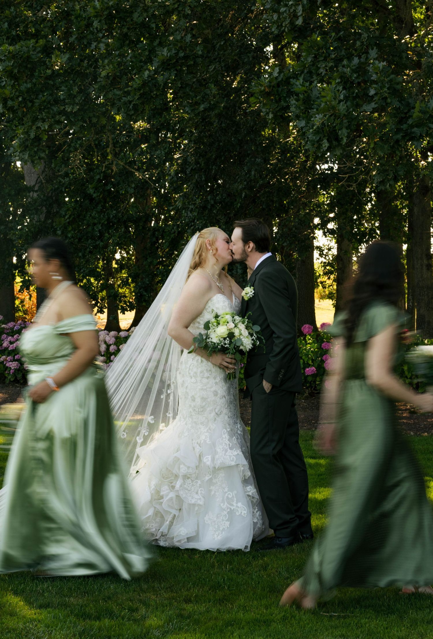 Bride and groom share a kiss outdoors while bridesmaids in sage green gowns walk past in a lush garden setting.