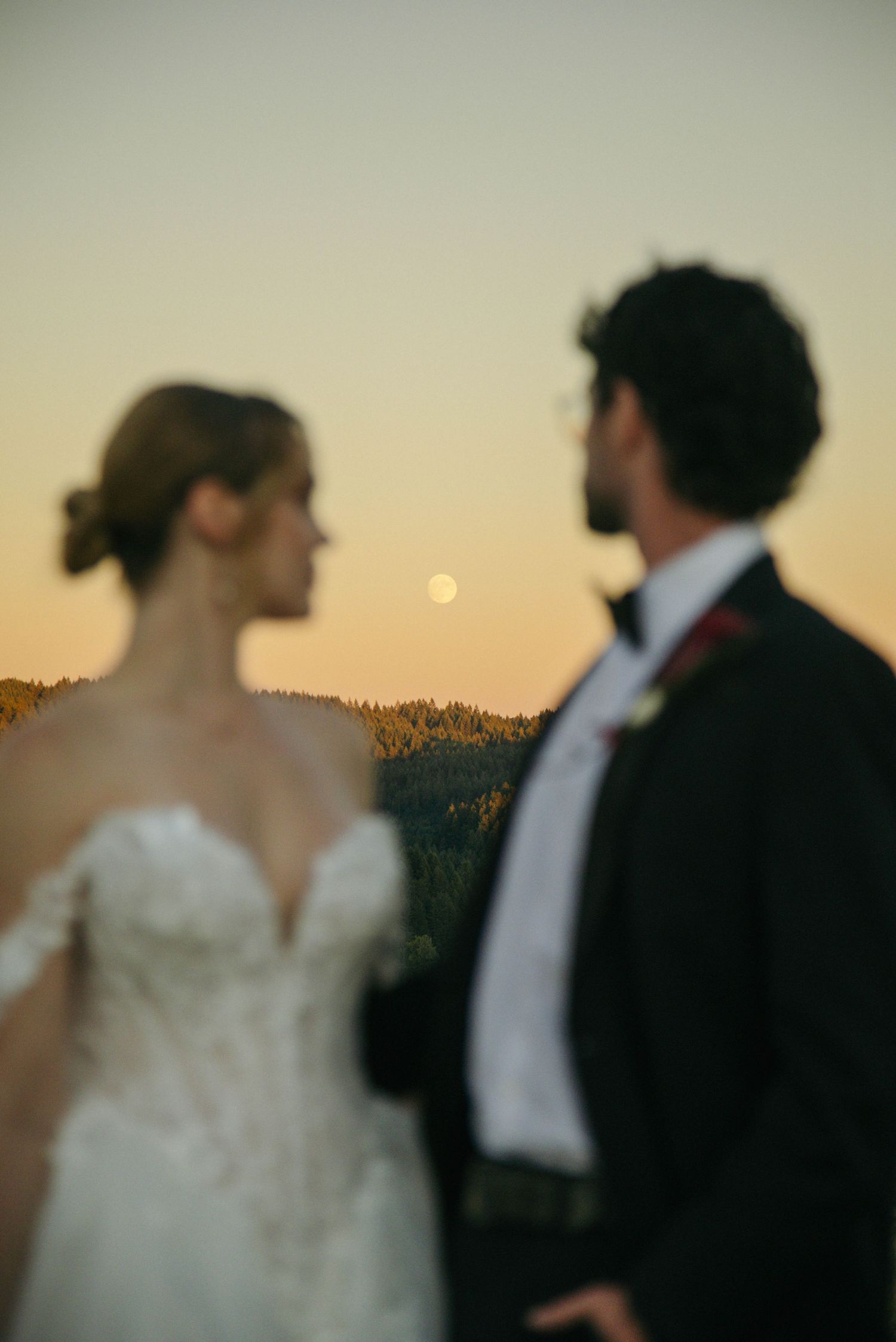 Bride and groom stand together at sunset with a full moon rising over forested hills in a romantic wedding portrait.