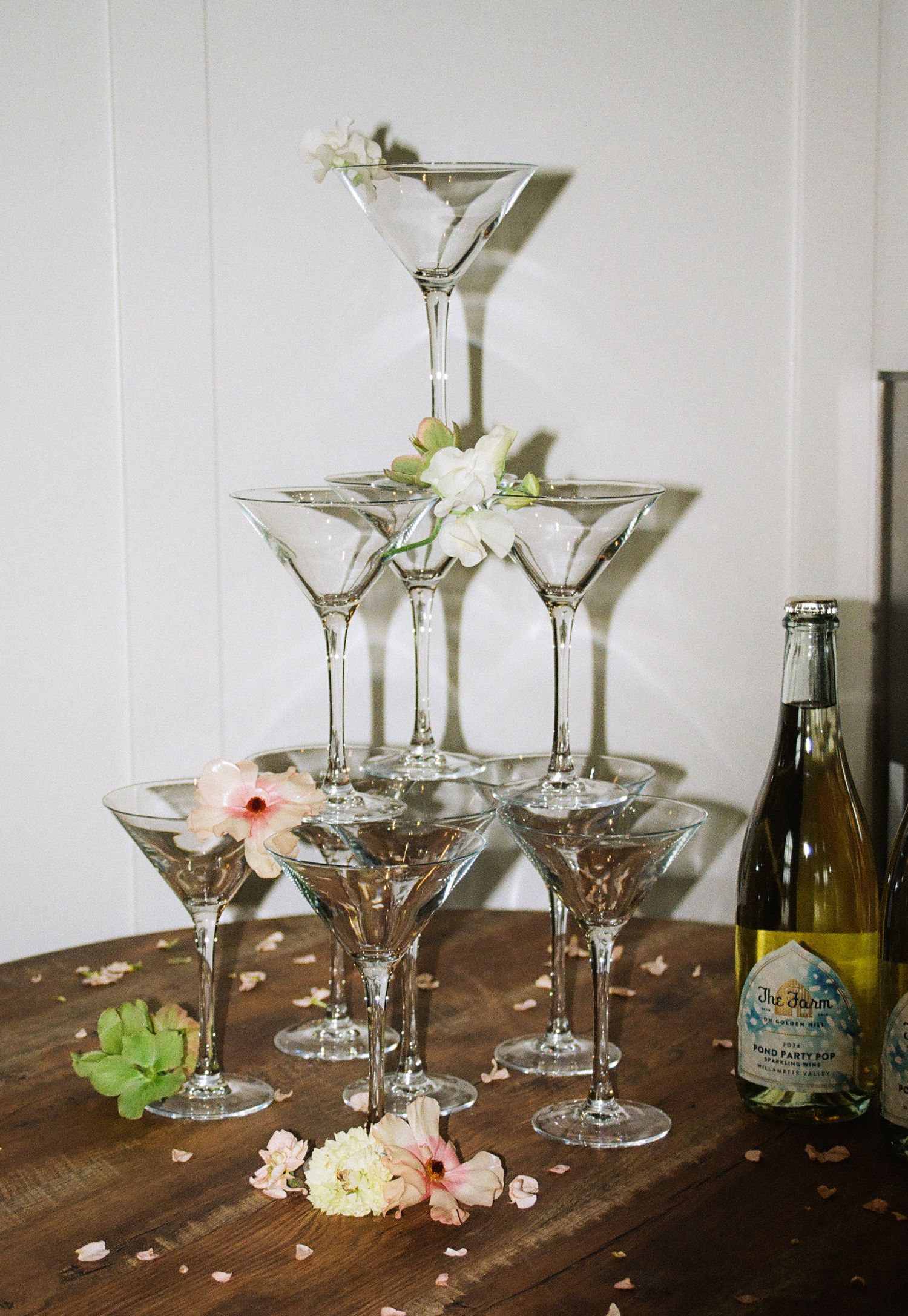 Elegant martini glass tower pyramid with floral accents beside a champagne bottle on a wooden table.