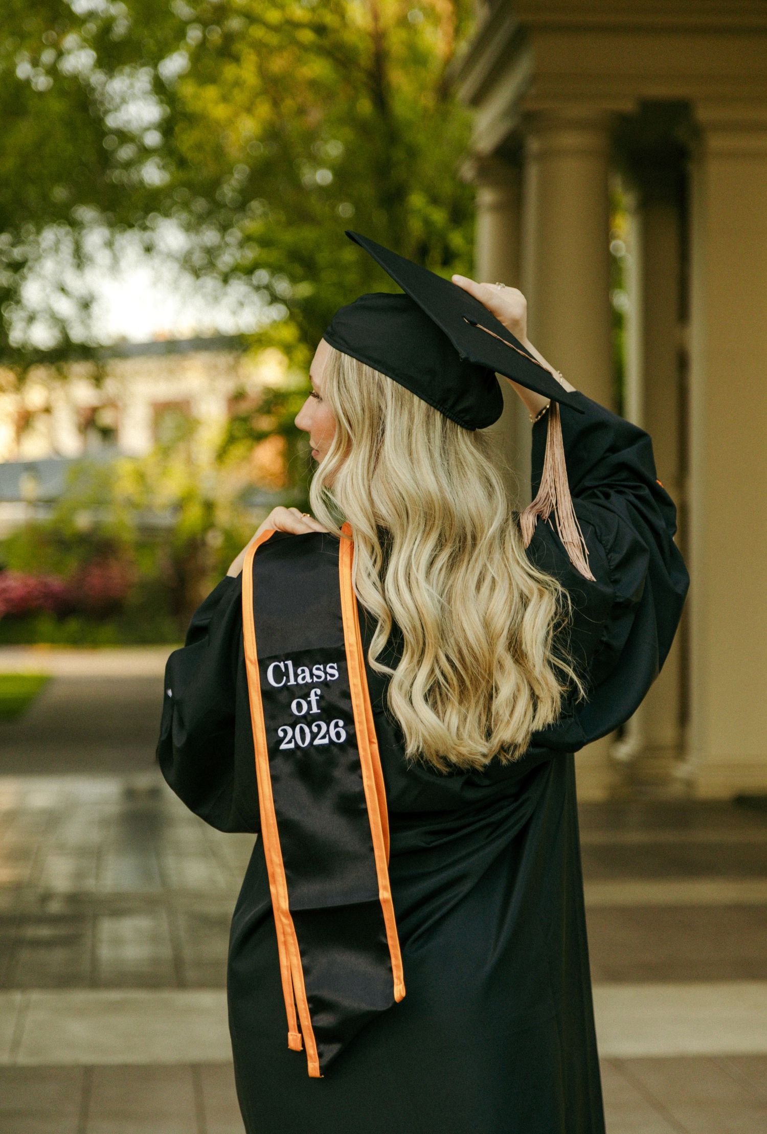 Blonde woman in black graduation gown and cap wearing a Class of 2026 stole with orange trim outdoors.