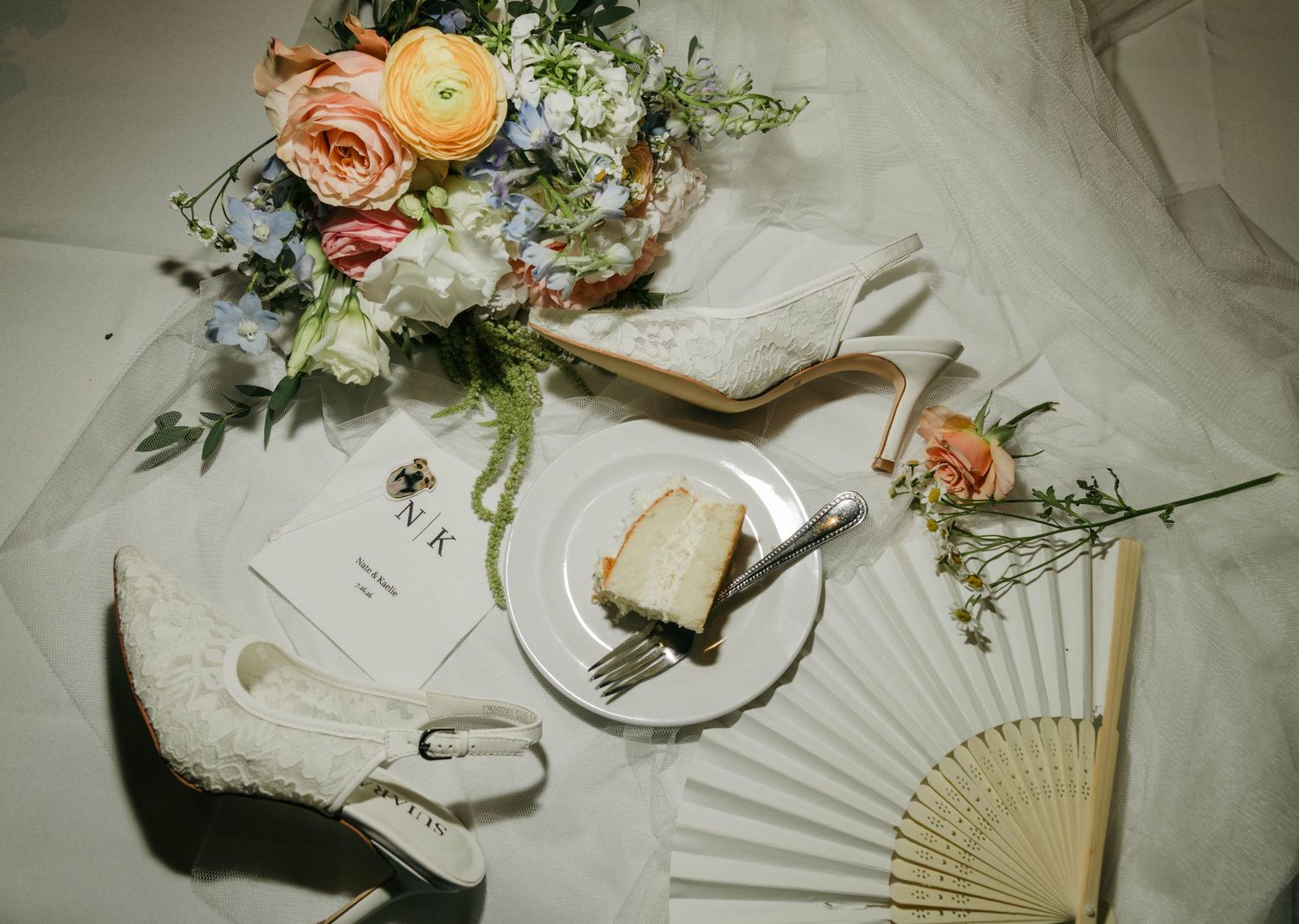 Elegant wedding flat lay with lace heels, floral bouquet, cake slice, hand fan, and stationery on white fabric.