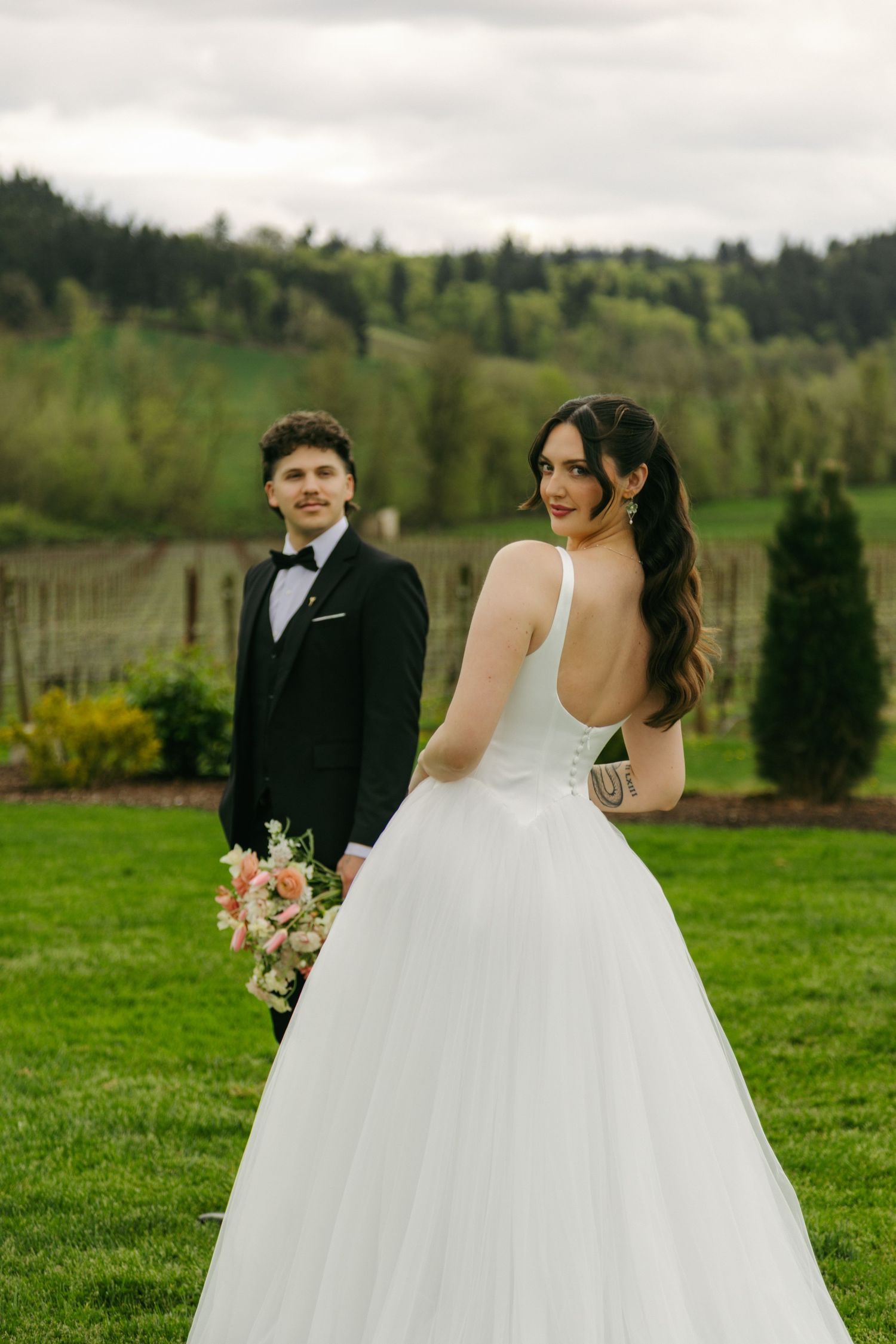 Bride in white backless gown holds pink bouquet, groom in black tuxedo stands behind on green lawn.