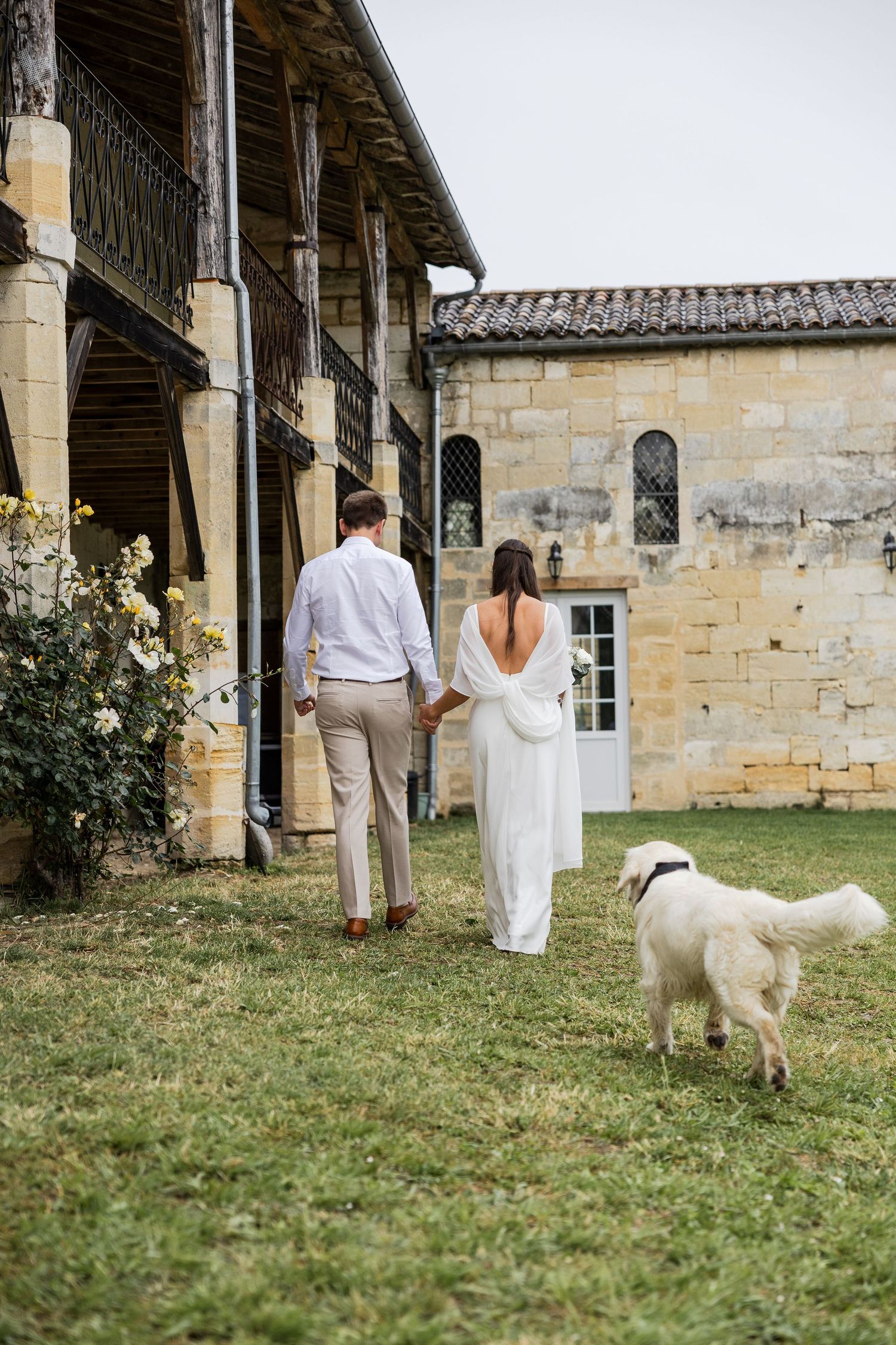 Bride and groom walking hand in hand with a white dog beside a rustic stone chateau on their wedding day.