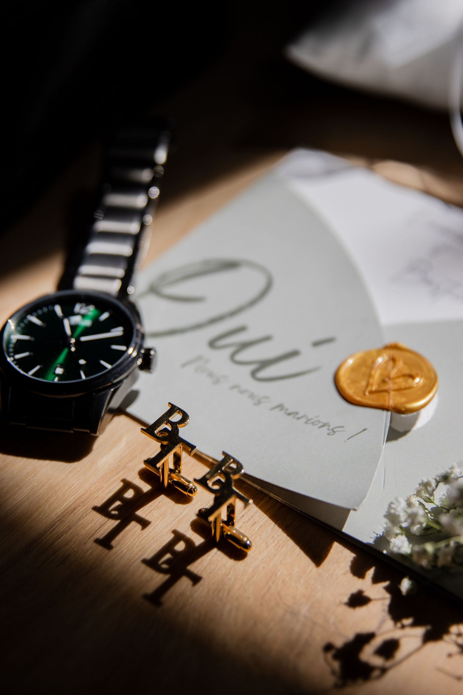 Elegant wedding flat lay with black watch, gold cufflinks, and a card reading Oui on a wooden surface.