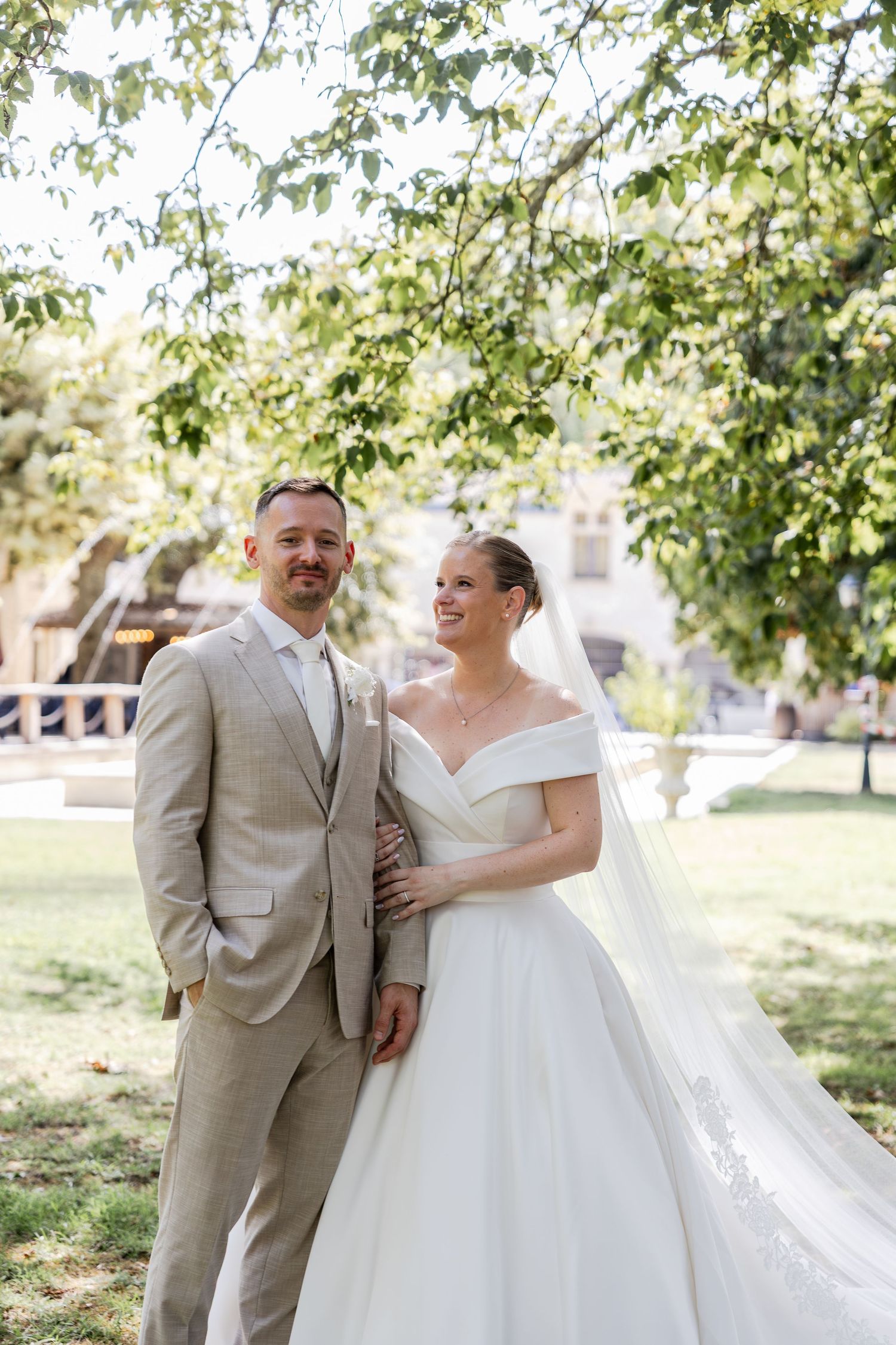 Bride in off-shoulder white gown and groom in tan suit pose together outdoors under green tree on wedding day.