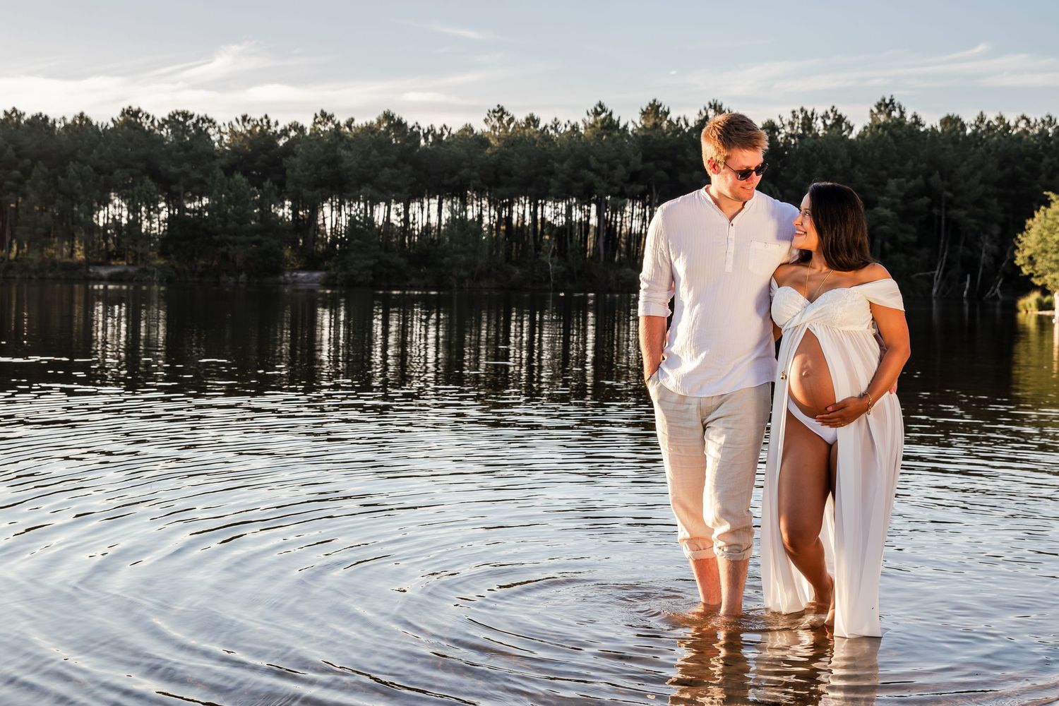 Pregnant couple in white outfits standing in a lake with trees reflected in the water during golden hour.
