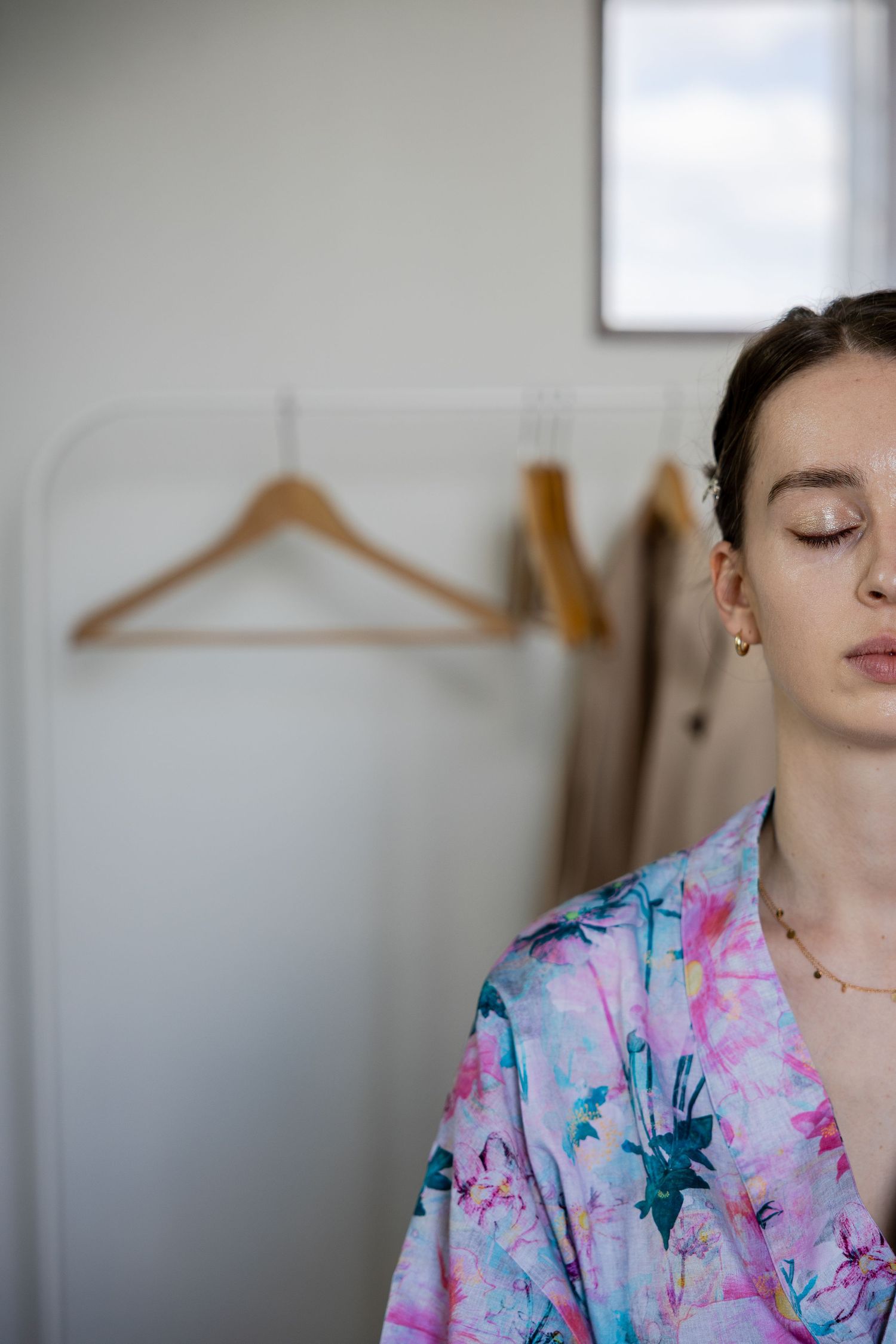 Young woman with closed eyes wearing a colorful floral kimono robe, standing in front of wooden clothes hangers.