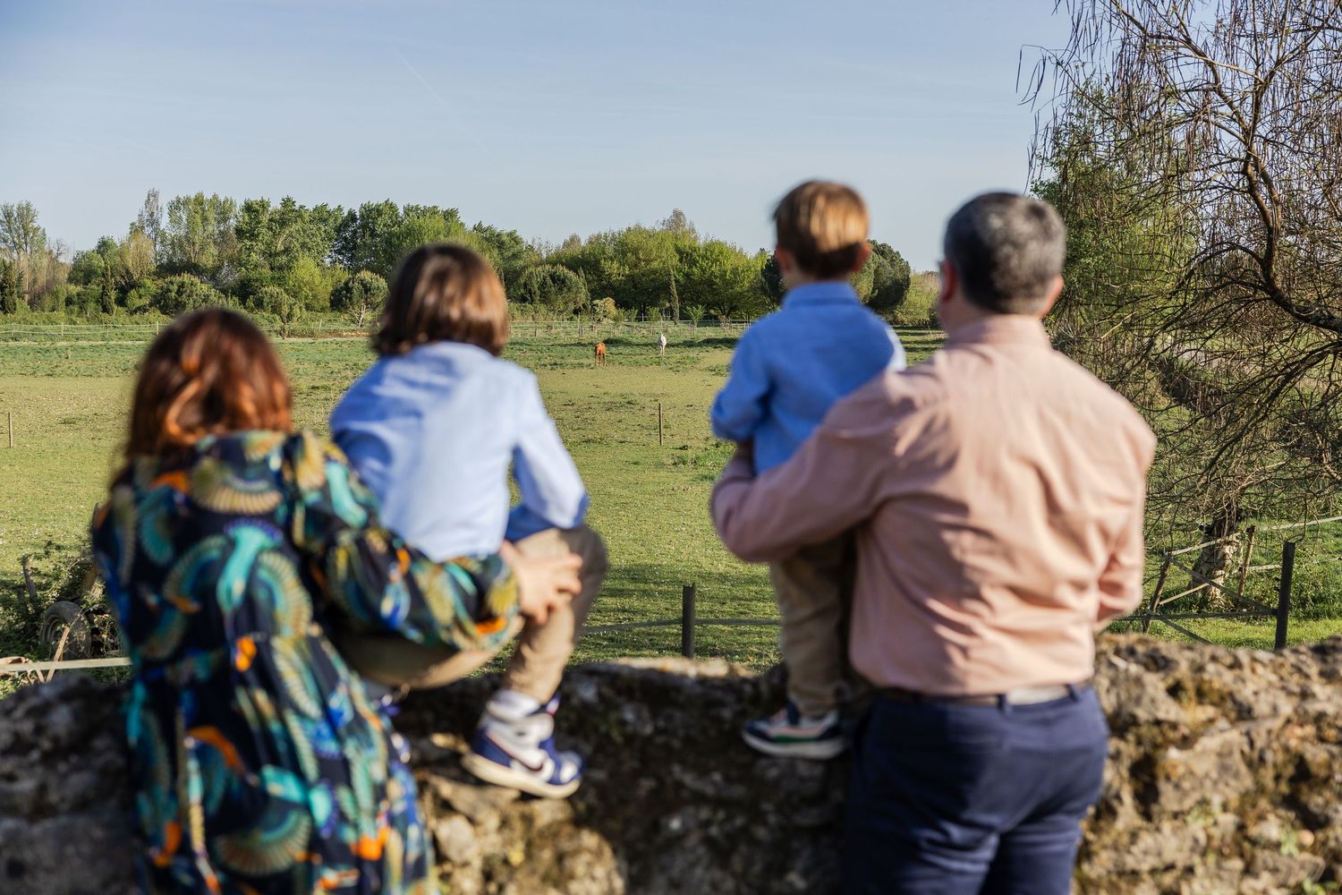 Family of four standing by a stone wall overlooking a lush green countryside on a sunny day.