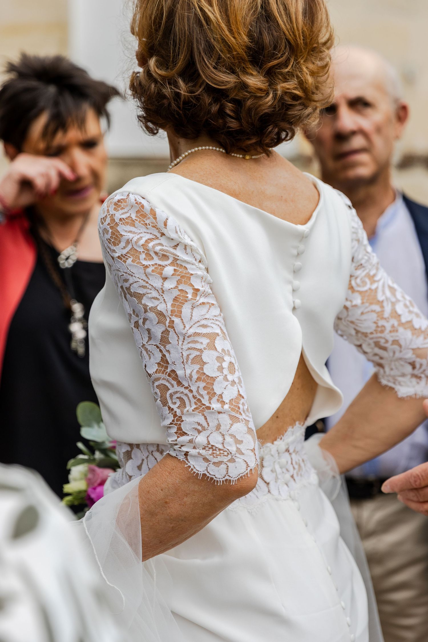 Bride in elegant white lace wedding dress with open back detail, standing with guests at outdoor ceremony.