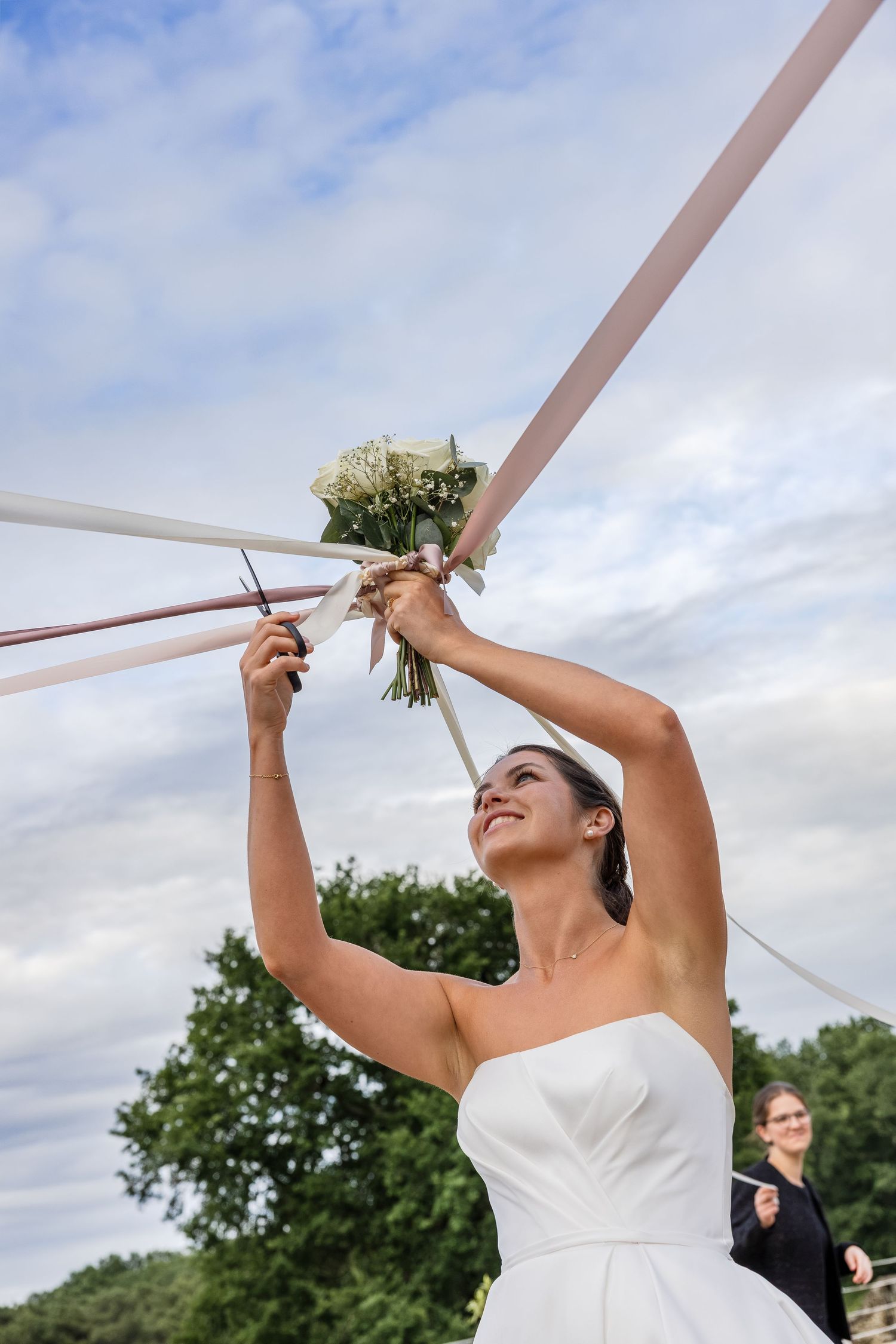 Smiling bride in white strapless gown holds floral bouquet with ribbons aloft outdoors on her wedding day.