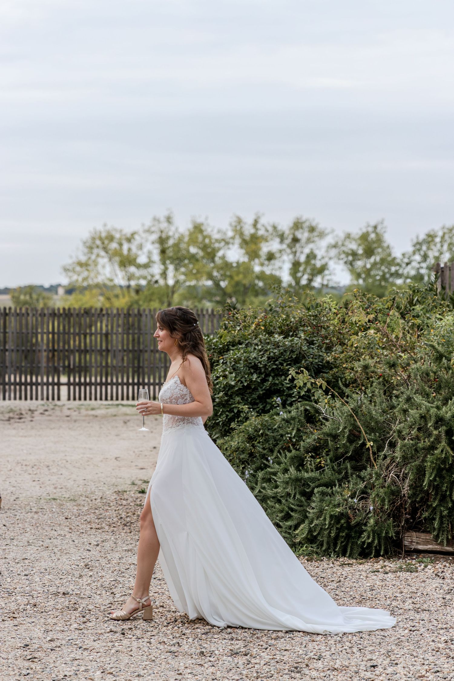 Bride in elegant white gown with lace bodice and slit skirt holding champagne outdoors near greenery and wooden fence.