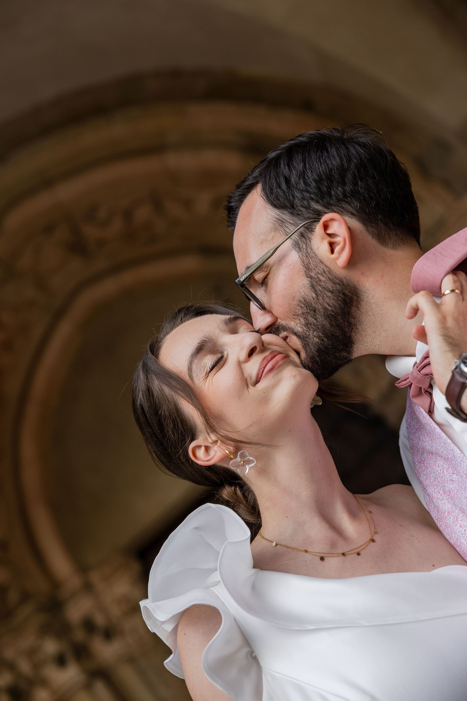 Groom with glasses and beard tenderly kissing smiling bride in white ruffled wedding dress indoors.
