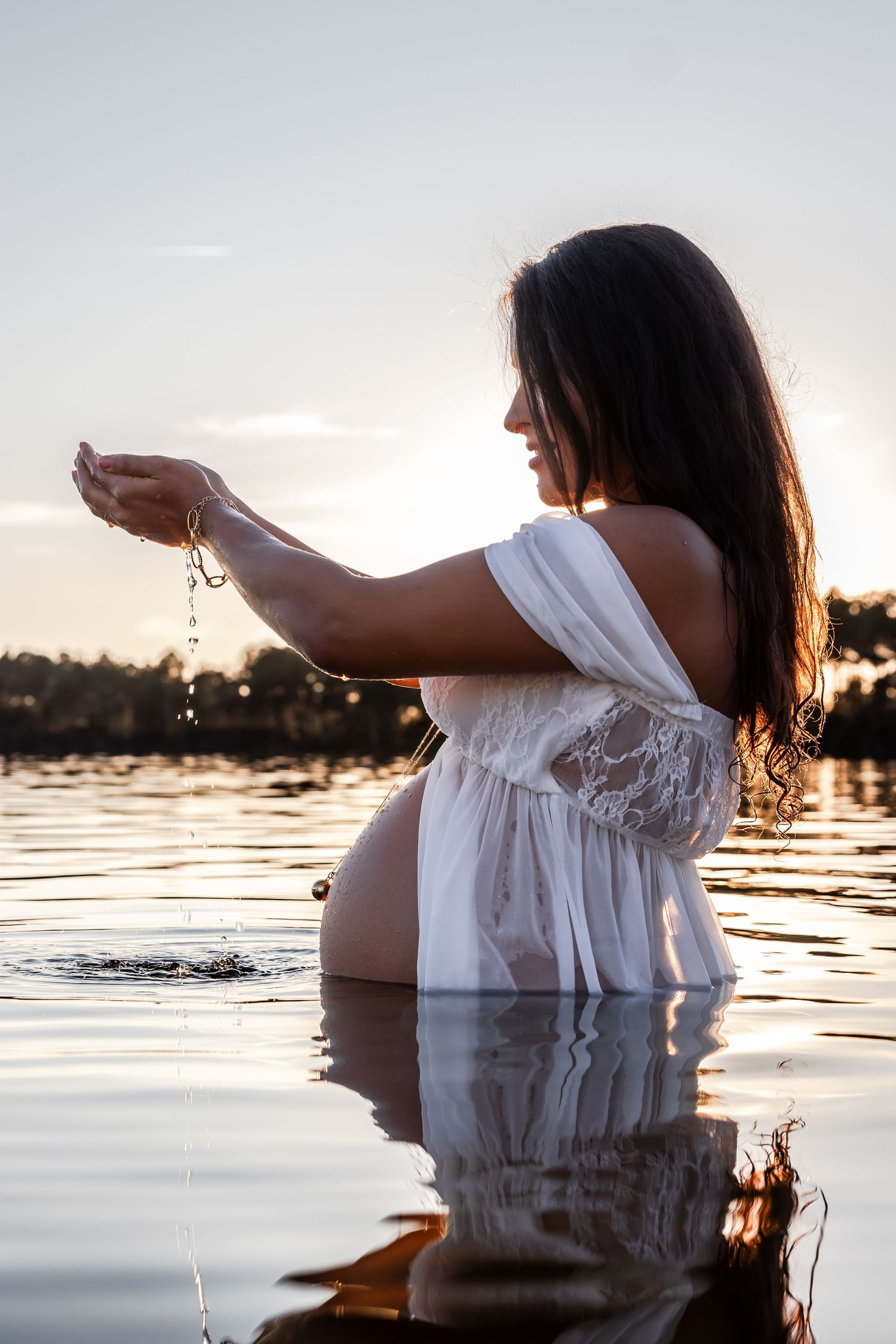 Woman in white dress standing in lake at sunset, letting water drip from her hands.