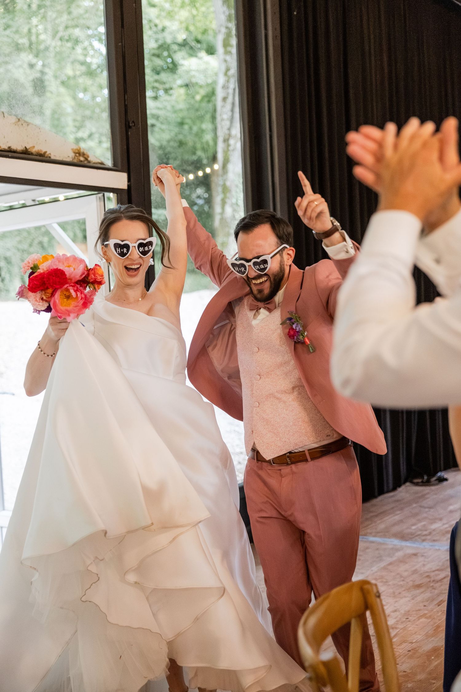 A joyful bride and groom wearing heart-shaped sunglasses celebrate their wedding reception entrance.
