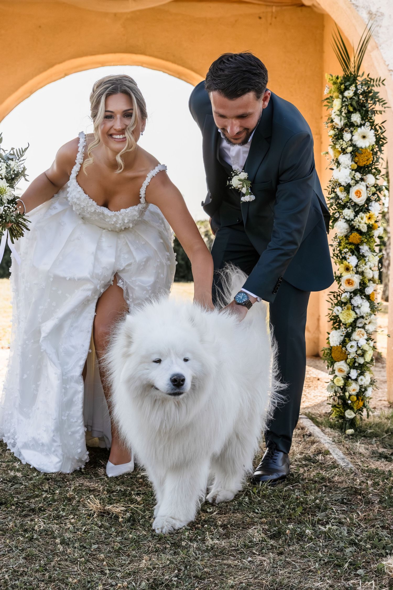 Bride and groom petting a fluffy white Samoyed dog at their outdoor wedding ceremony with floral decorations.