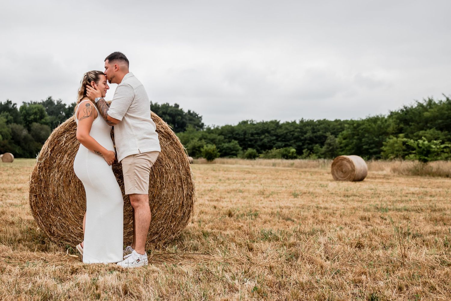 Couple embracing near a hay bale in a golden field, woman in white dress, man in beige outfit, cloudy sky backdrop.