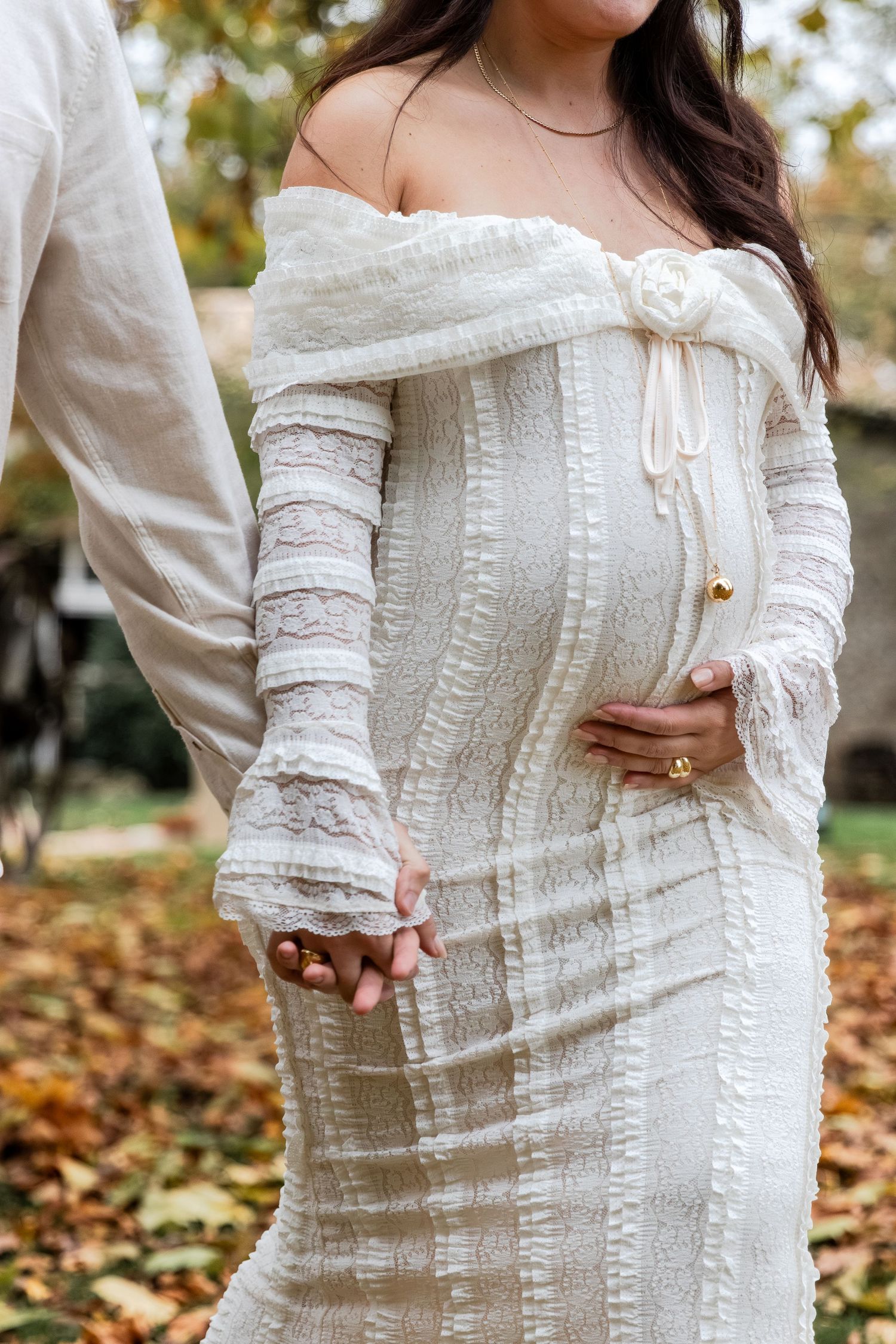 Pregnant woman in white off-shoulder lace dress holding hands with partner outdoors among autumn leaves.