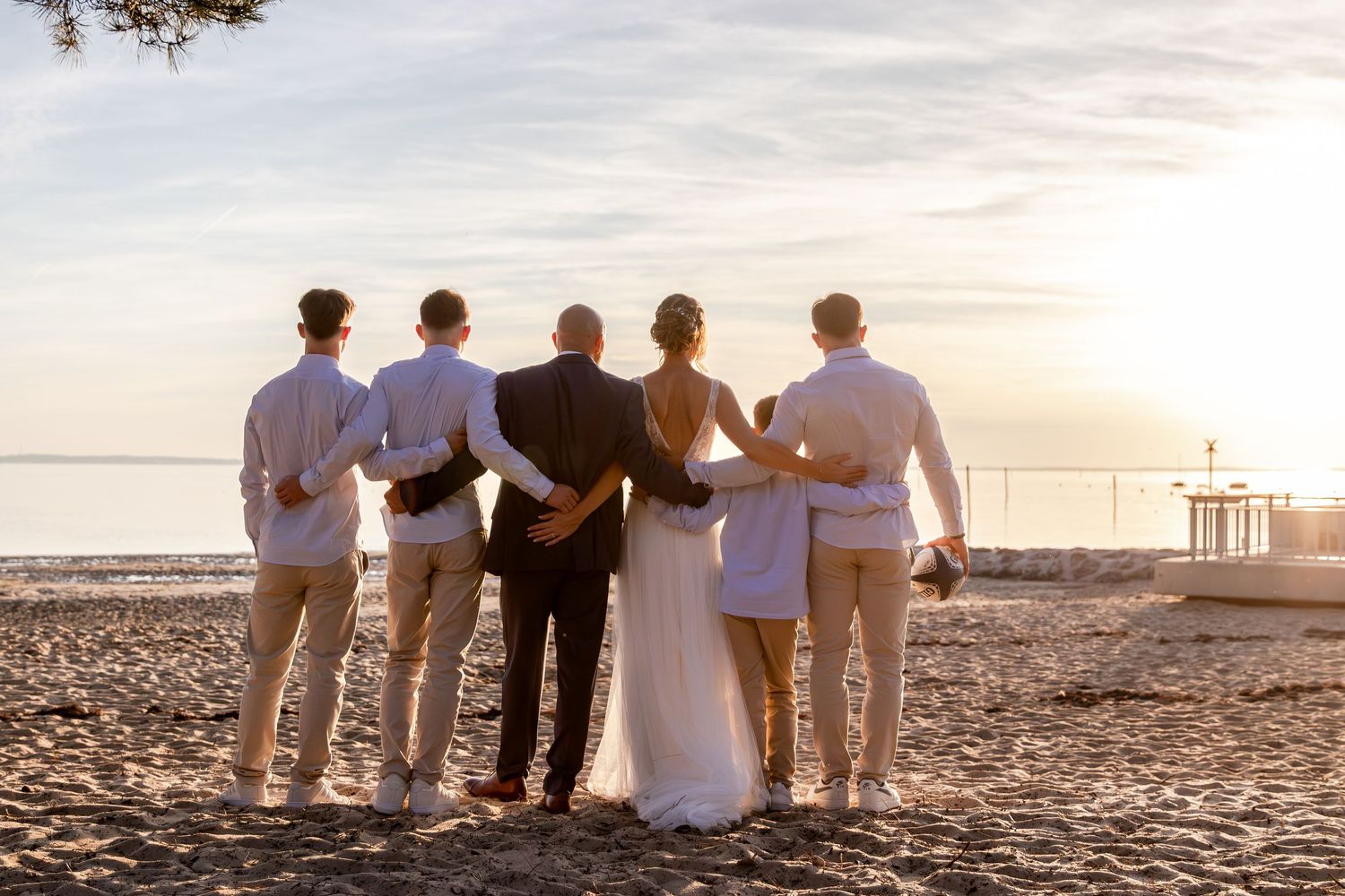 Wedding party in formal attire standing together on a sandy beach at sunset, viewed from behind.