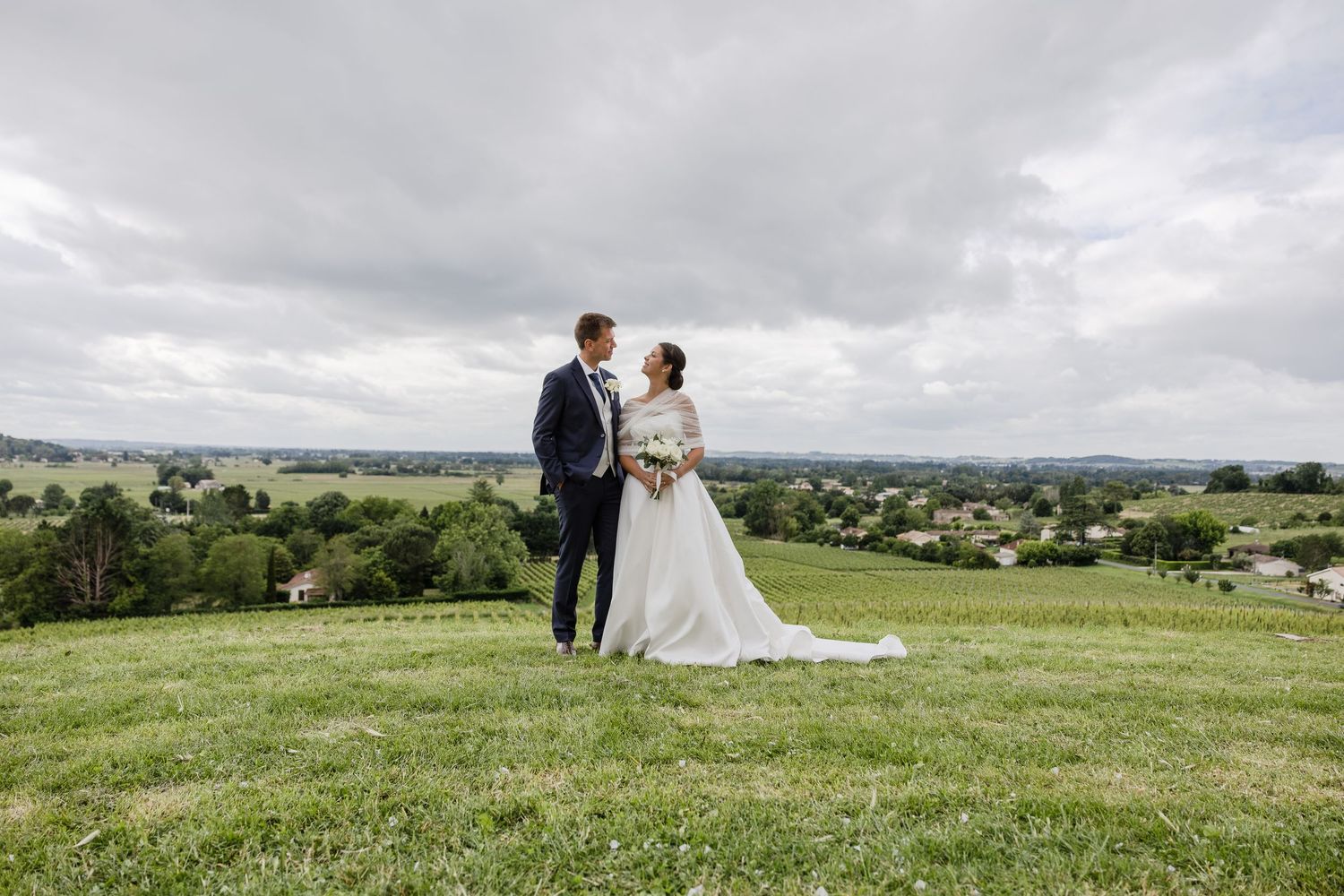 Bride and groom share a romantic moment on a green hillside with a scenic countryside view under cloudy skies.
