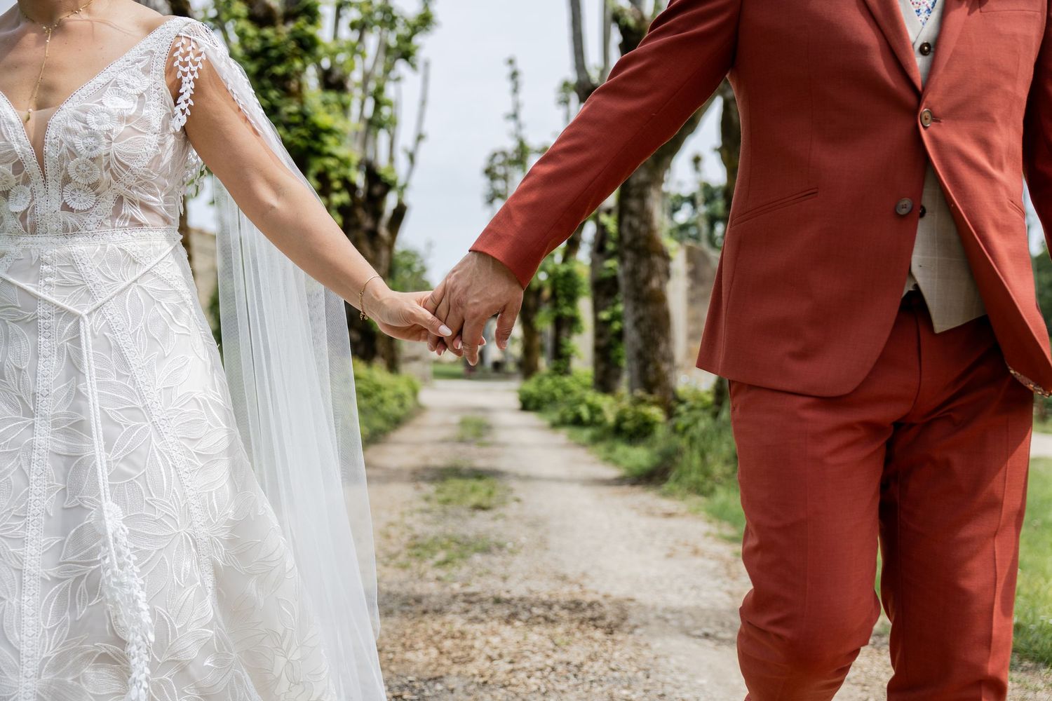 Bride and groom holding hands in lace wedding dress and rust suit on a tree-lined garden path.