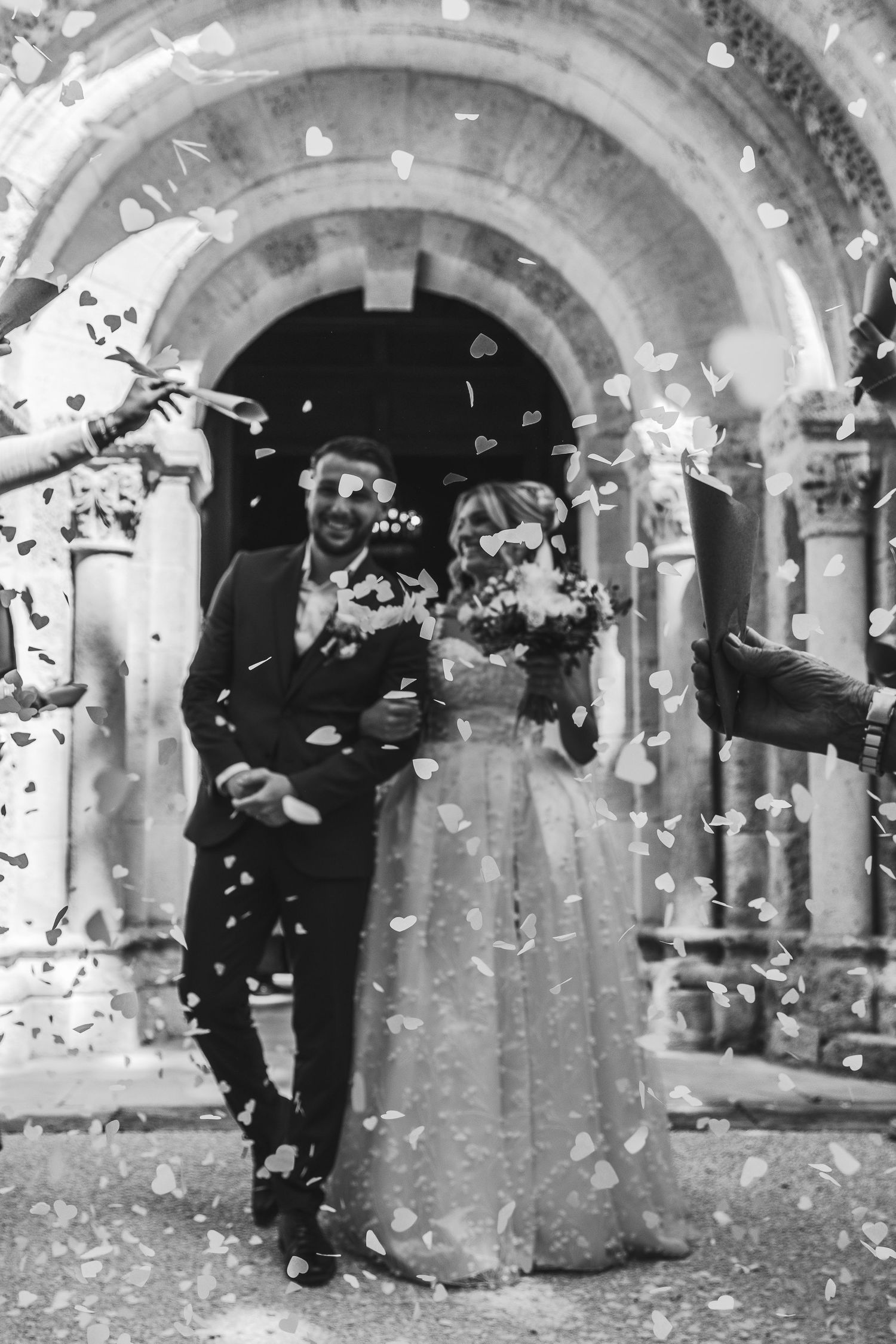 Black and white photo of newlyweds leaving church amid confetti shower, bride holding bouquet.
