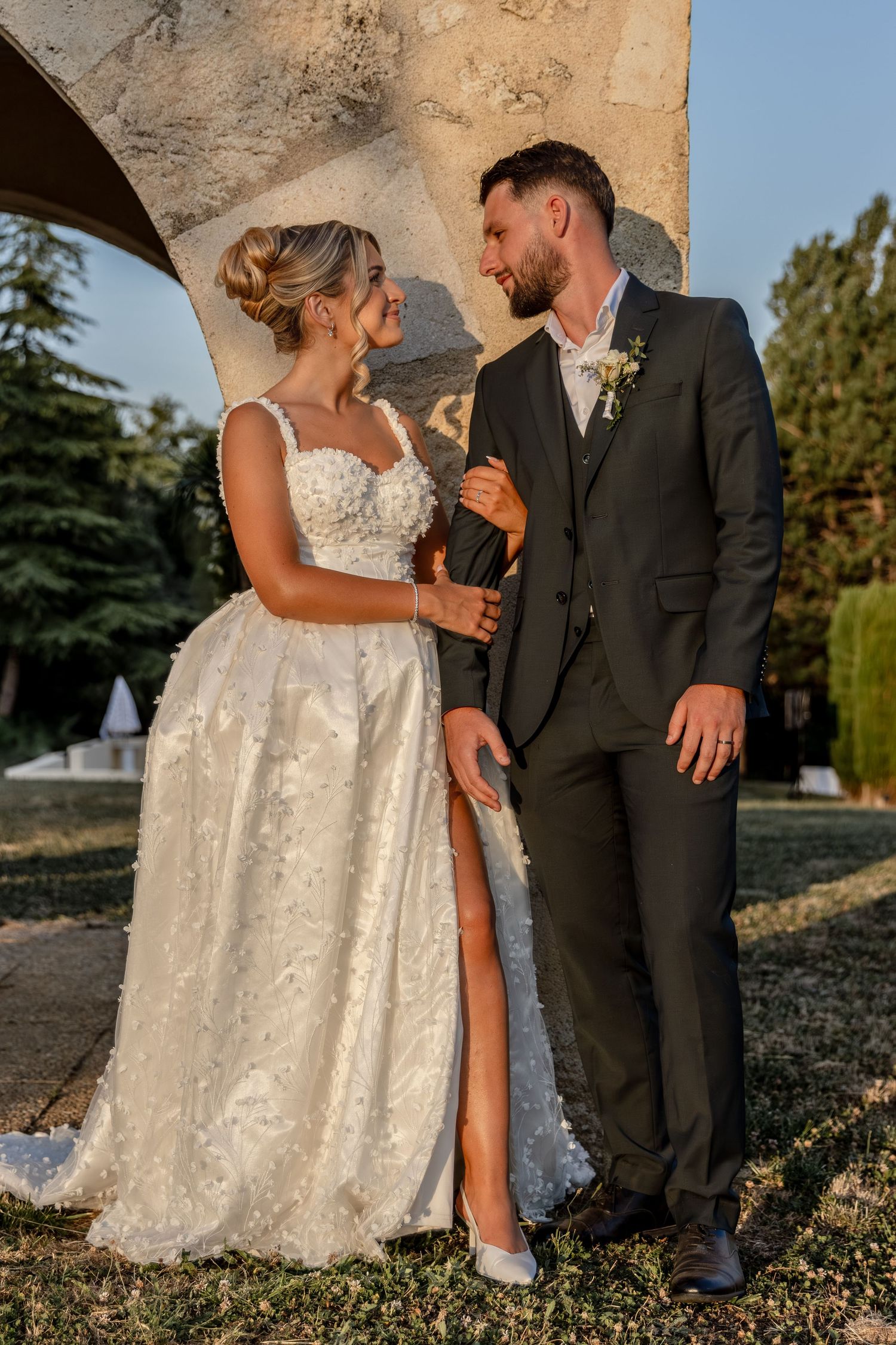 A bride and groom share a romantic moment outdoors, she in a floral white gown, he in a dark suit with boutonniere.