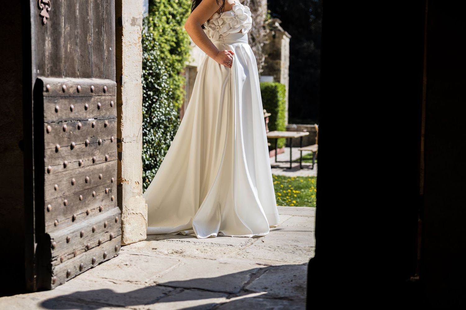 Bride in elegant white wedding gown standing in sunlit courtyard doorway, flowing skirt and lace bodice visible.