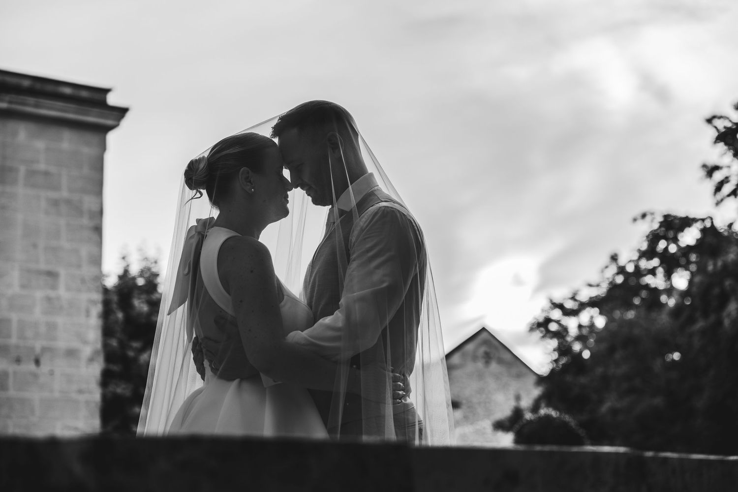 Black and white wedding photo of bride and groom in romantic embrace, veil flowing, with historic building backdrop.