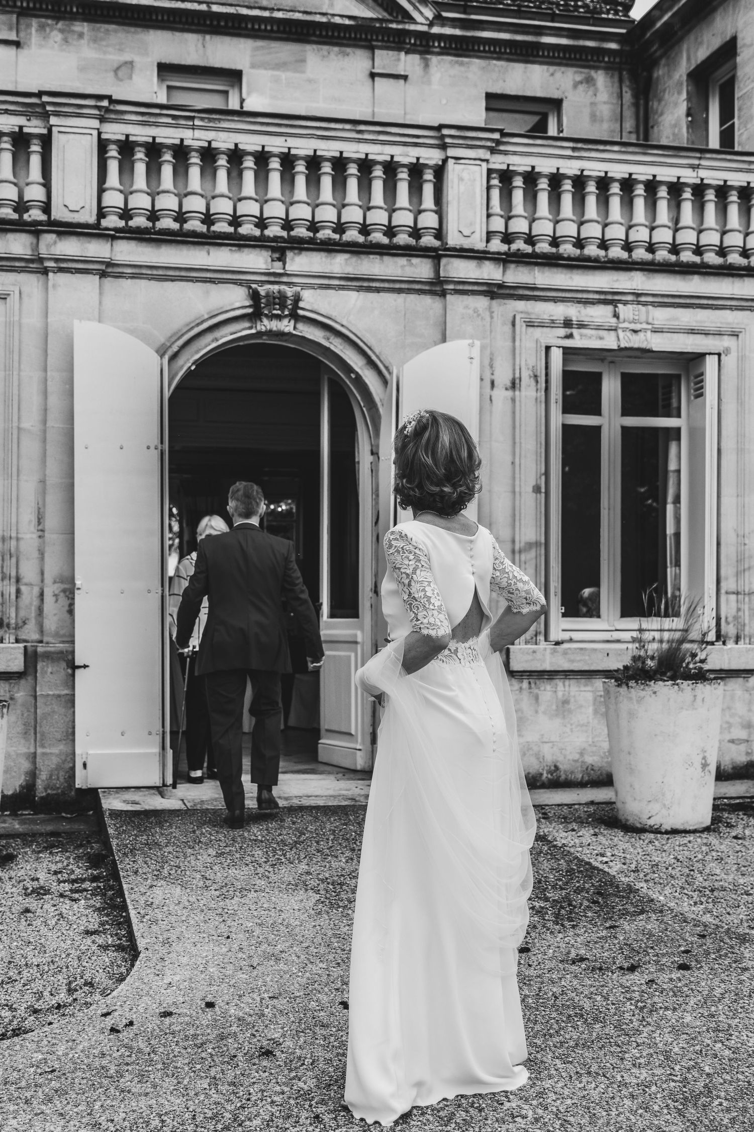 Black and white photo of a bride in a lace-back gown watching a couple enter an elegant French chateau.