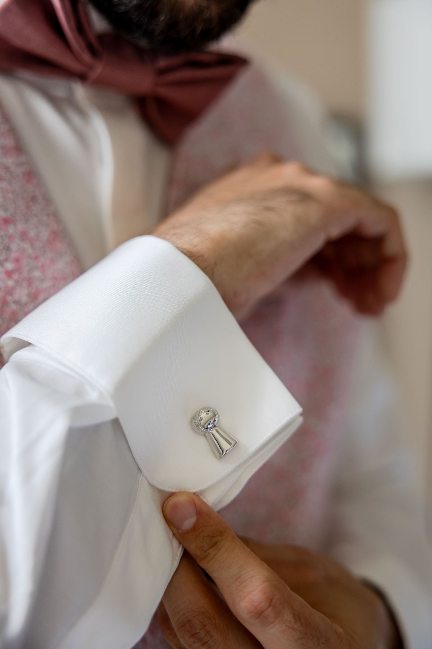 Close-up of a silver cufflink on a white dress shirt sleeve, with a man in a pink bow tie in the background.