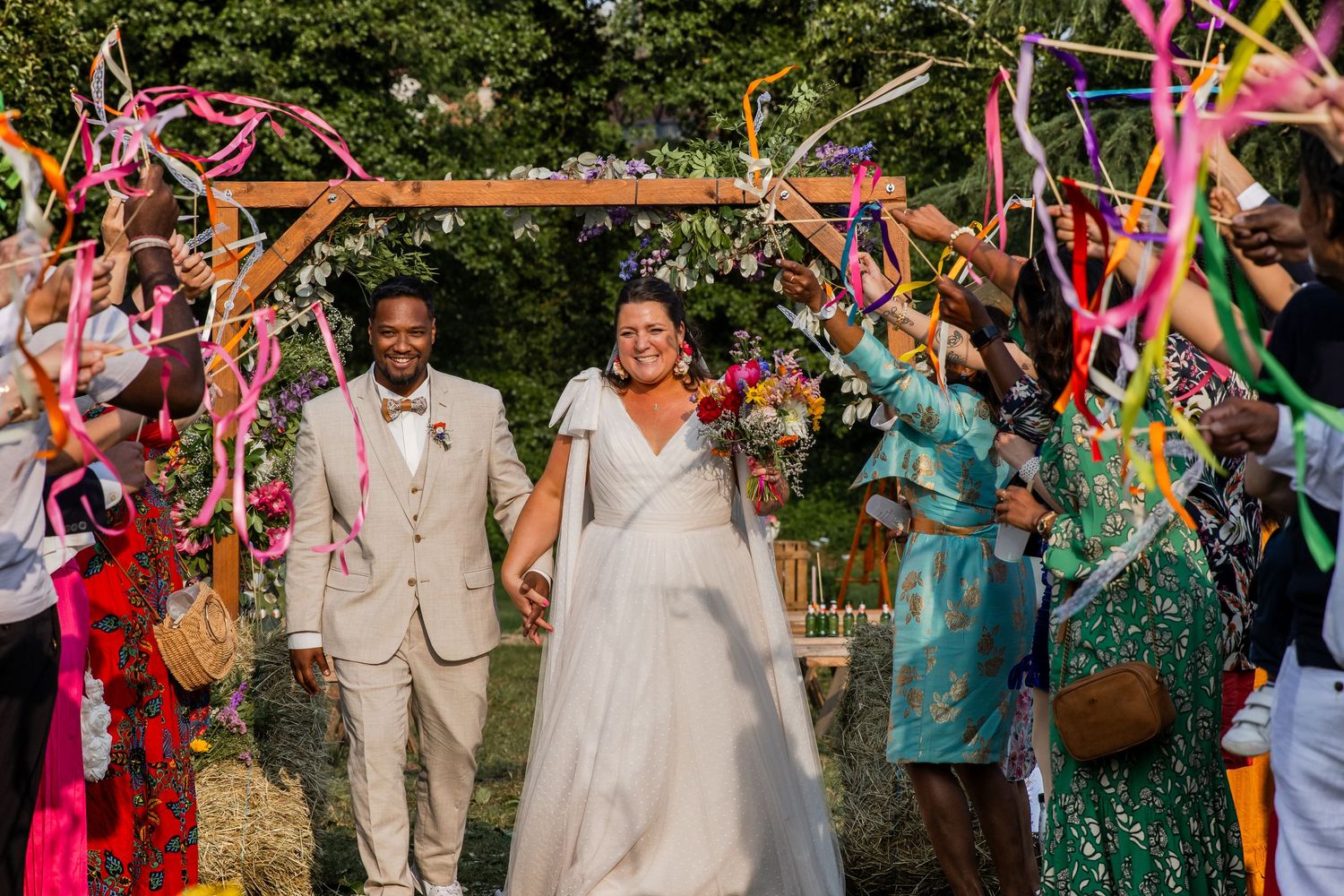 Joyful newlyweds walk under colorful ribbon arch at outdoor wedding ceremony with guests celebrating around them.