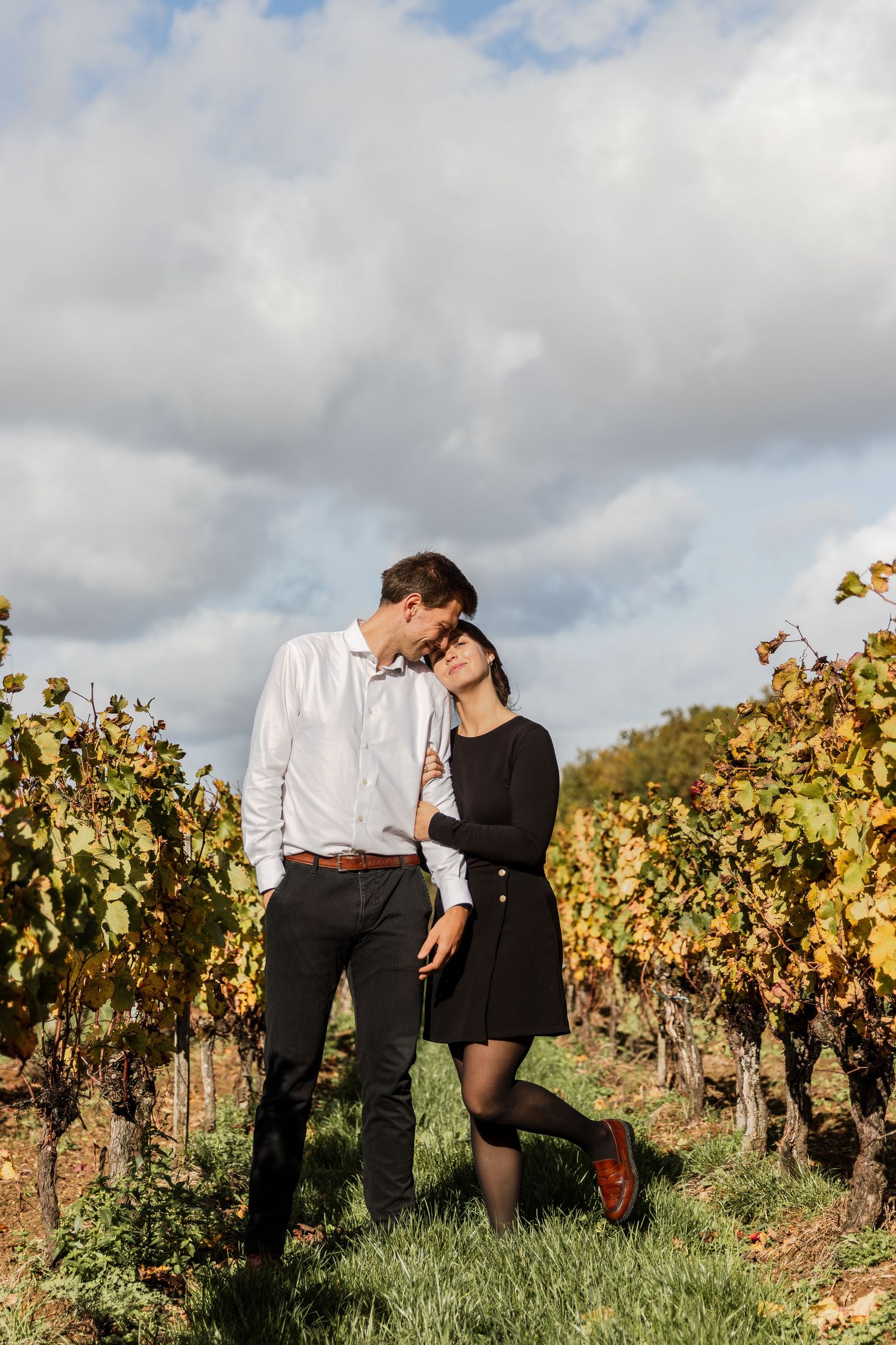 Romantic couple embracing in a vineyard during autumn, with golden foliage and cloudy sky in the background.
