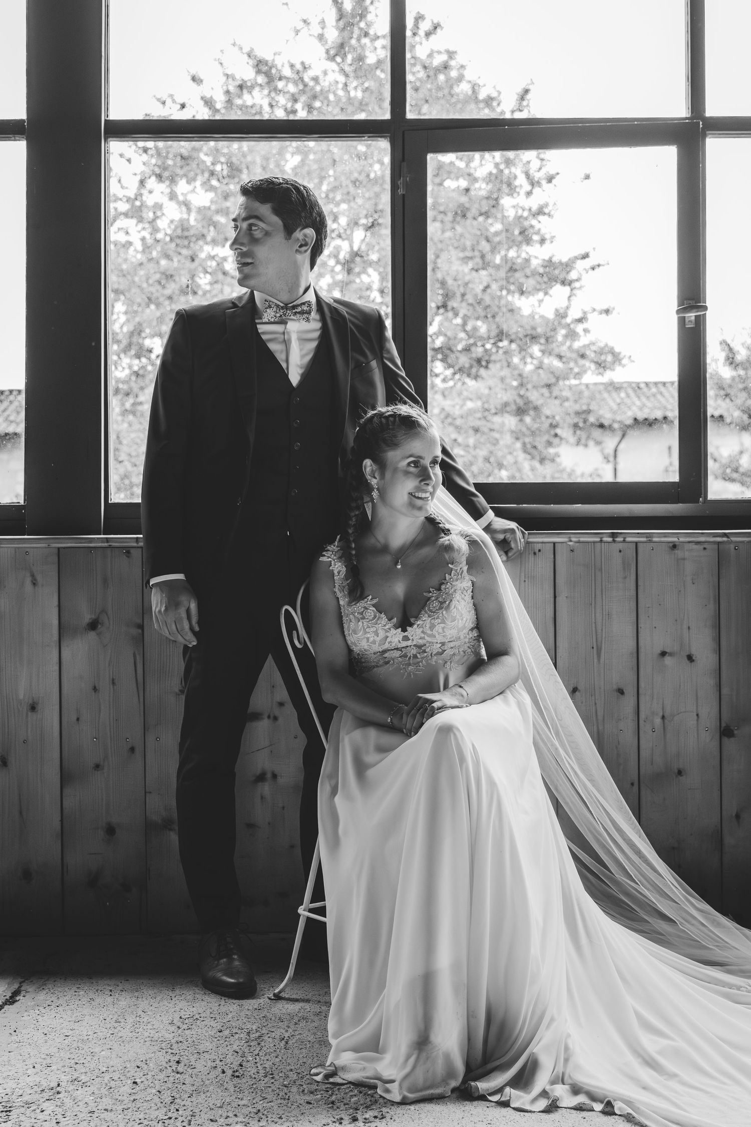 Black and white wedding photo of bride seated in gown with veil and groom standing by large window.