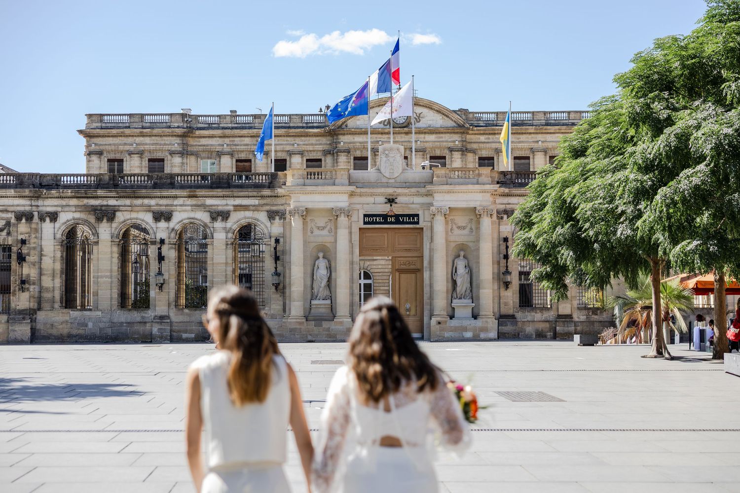 Two brides in white dresses walk toward a grand French government building with flags in Bordeaux.