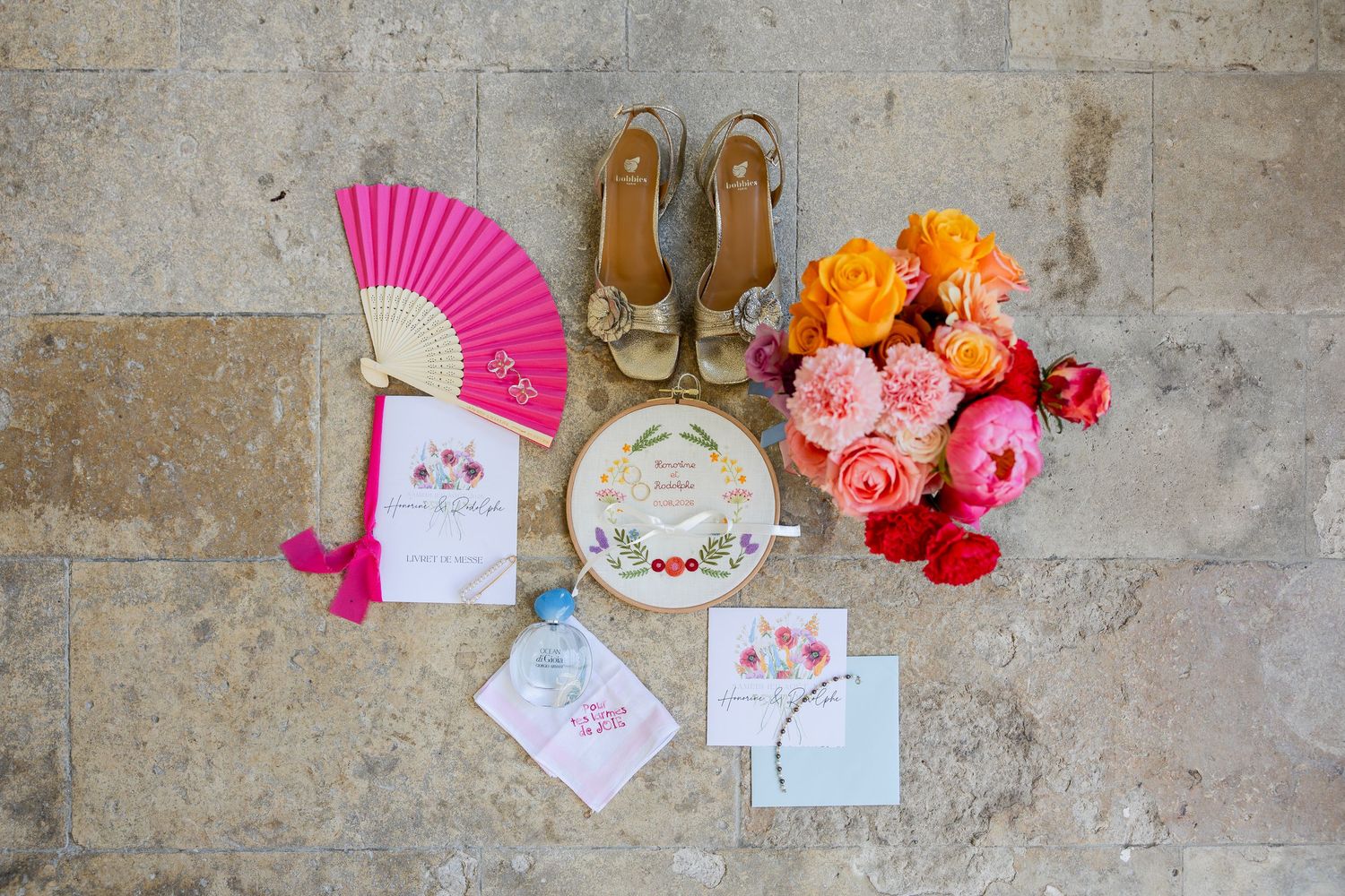 Colorful wedding flat lay with pink fan, gold heels, floral bouquet, invitation suite, and embroidered hoop on stone surface.