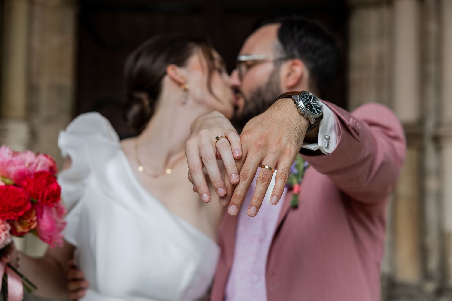 Newlyweds kissing while showing off wedding rings, bride holding pink bouquet, groom in pink suit outside church.
