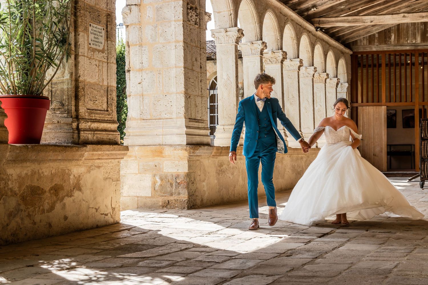 Bride and groom hold hands walking through sunlit stone archway colonnade in elegant wedding attire.