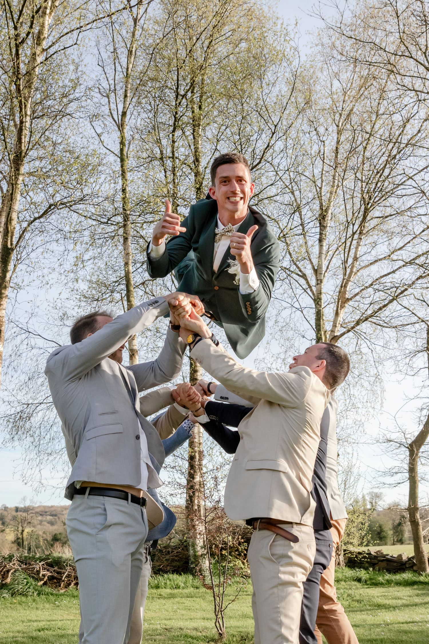 Groomsmen tossing groom in the air outdoors, celebrating wedding day with joy and laughter in a sunny garden setting.
