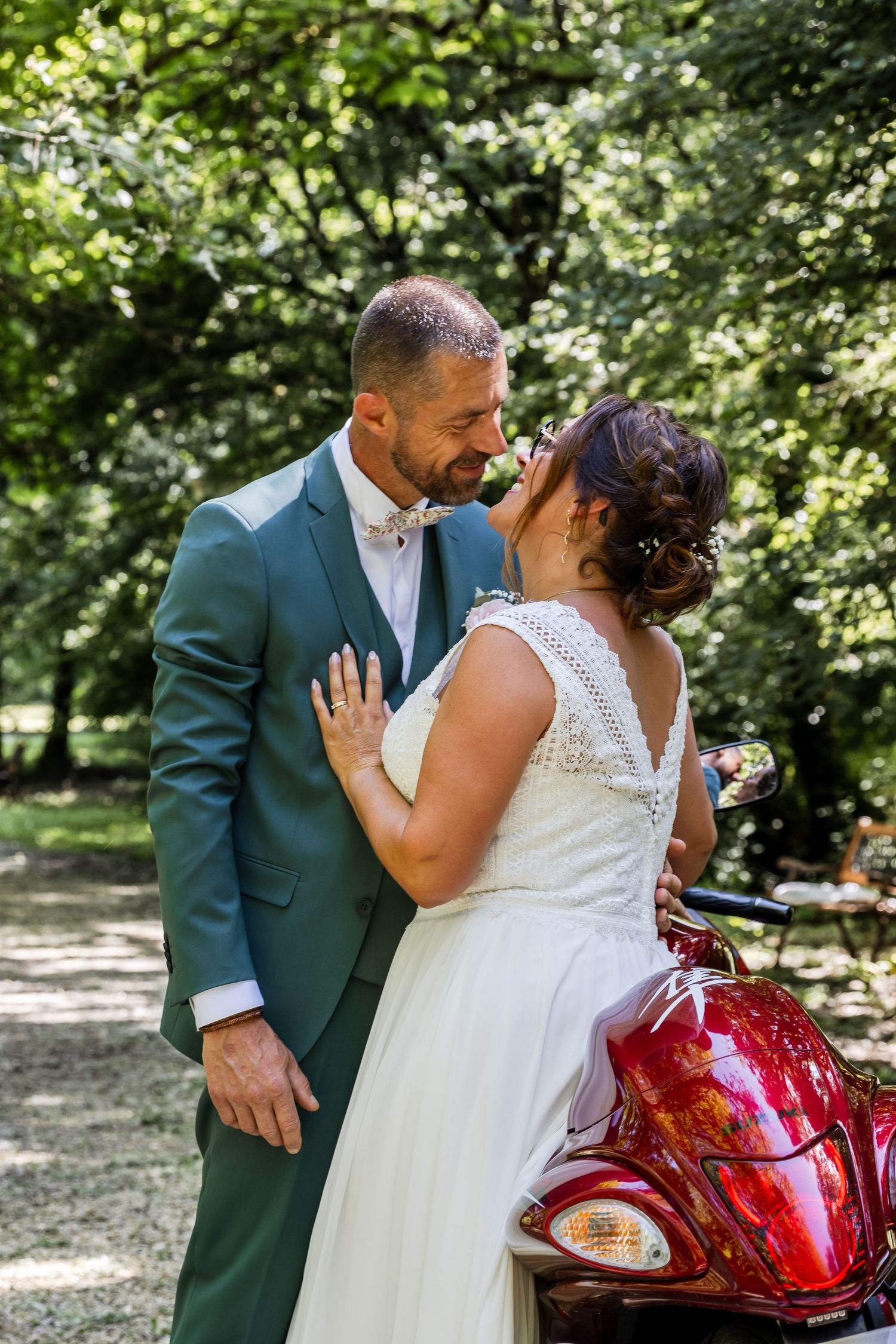 Bride and groom share a romantic moment near a red motorcycle in a lush green park setting.
