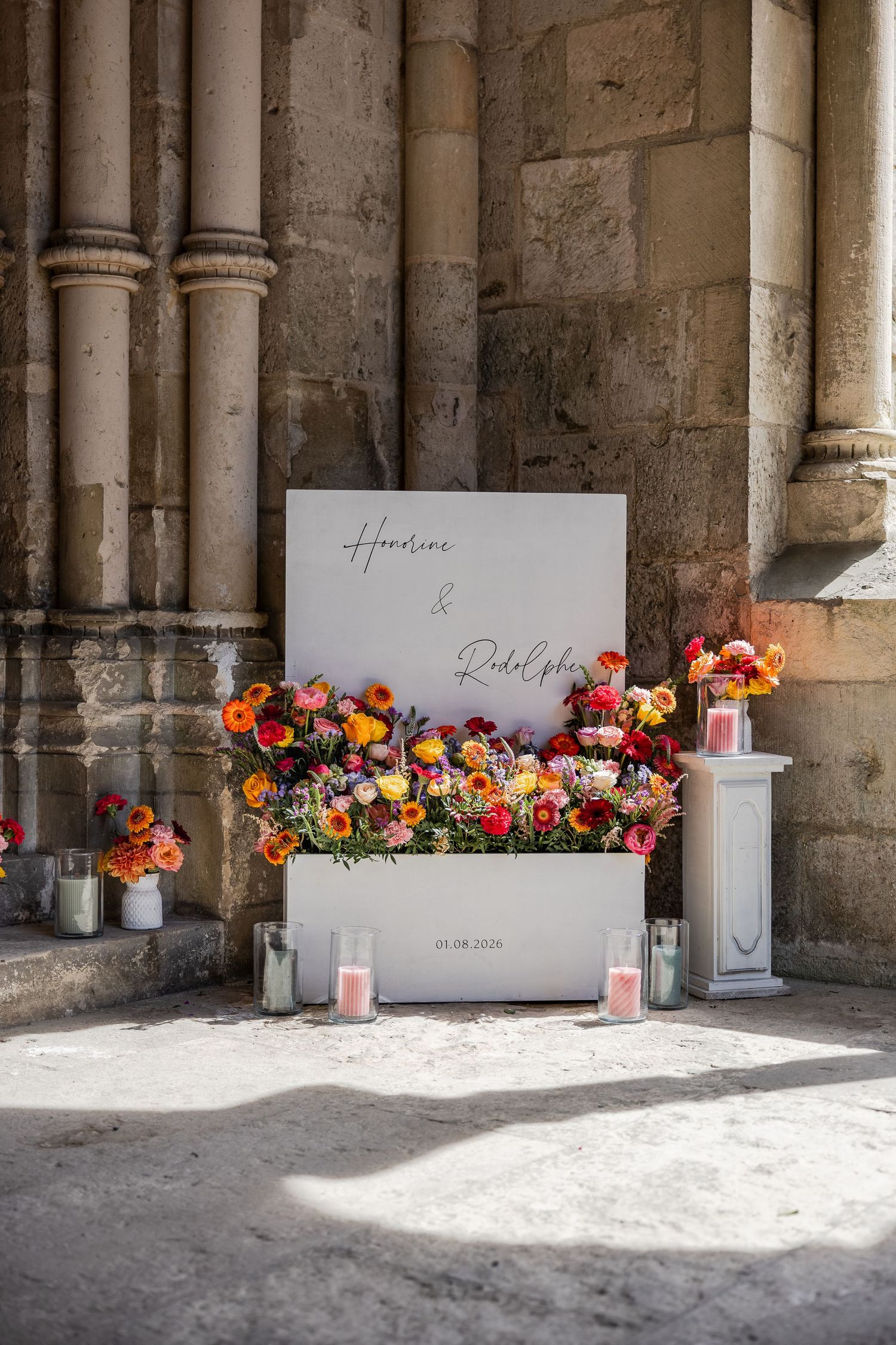 Elegant wedding welcome sign with colorful ranunculus flowers, candles, and couple names displayed at a stone church entrance.