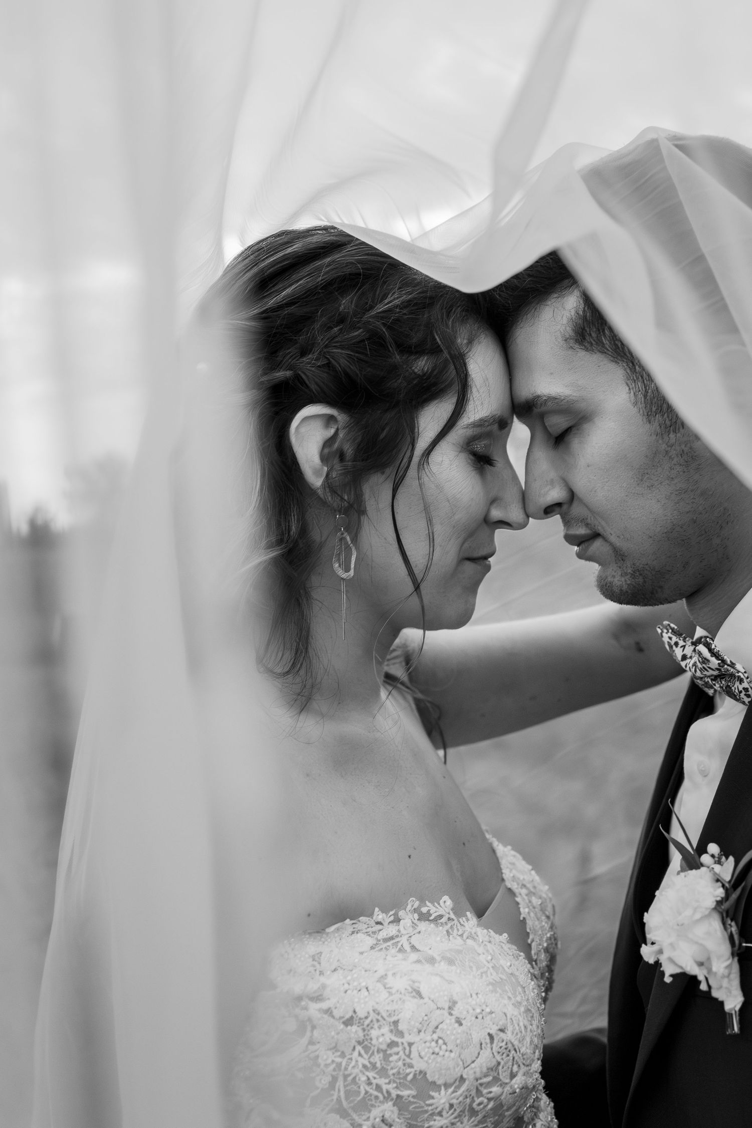 Black and white wedding photo of bride and groom touching foreheads tenderly under a flowing veil.