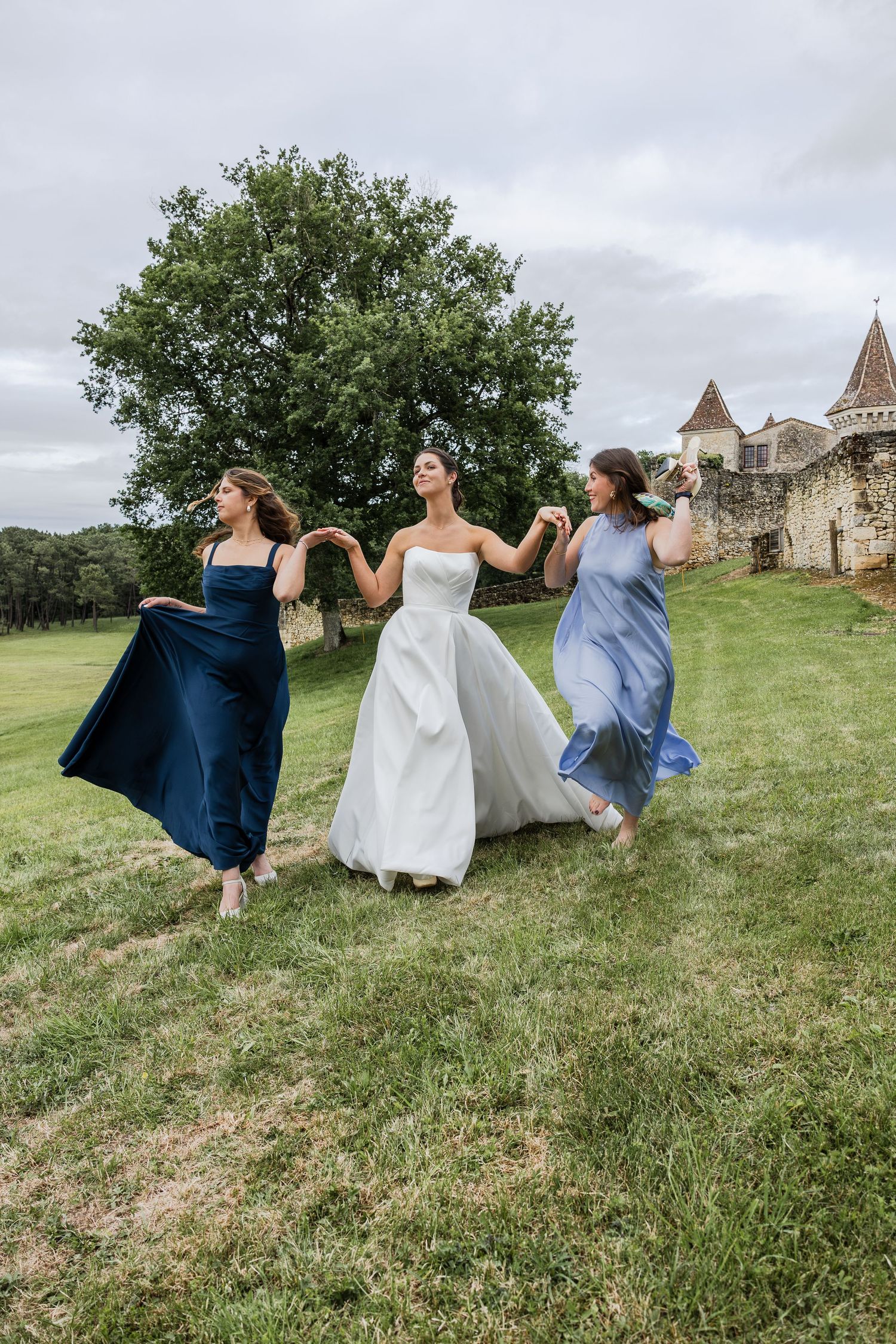 Bride in white gown runs hand-in-hand with two bridesmaids in blue dresses across green lawn near historic castle.