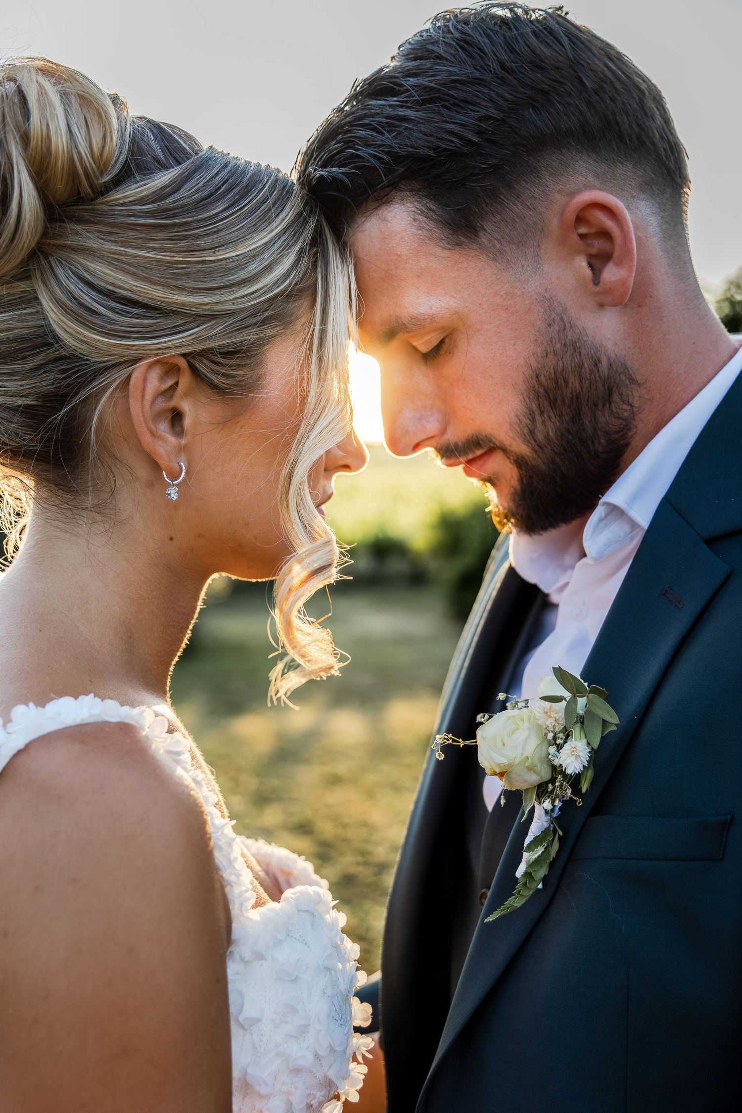 Bride and groom touching foreheads in golden sunset light, groom wearing boutonniere, intimate wedding portrait.