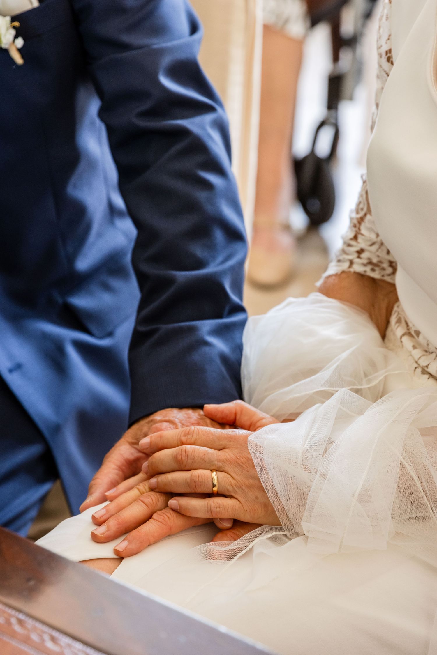 Bride and groom holding hands with wedding rings visible during a church ceremony, wearing white dress and blue suit.