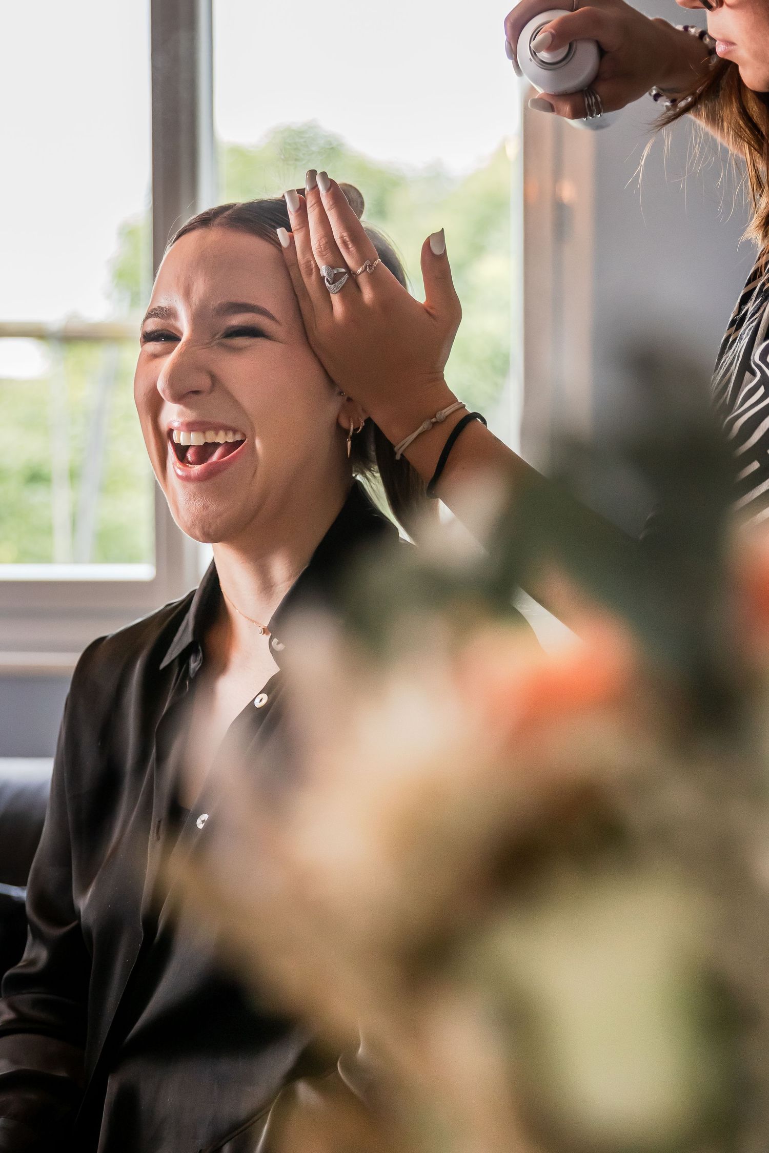 A woman laughs joyfully while a hairstylist applies hairspray during a bridal preparation session.