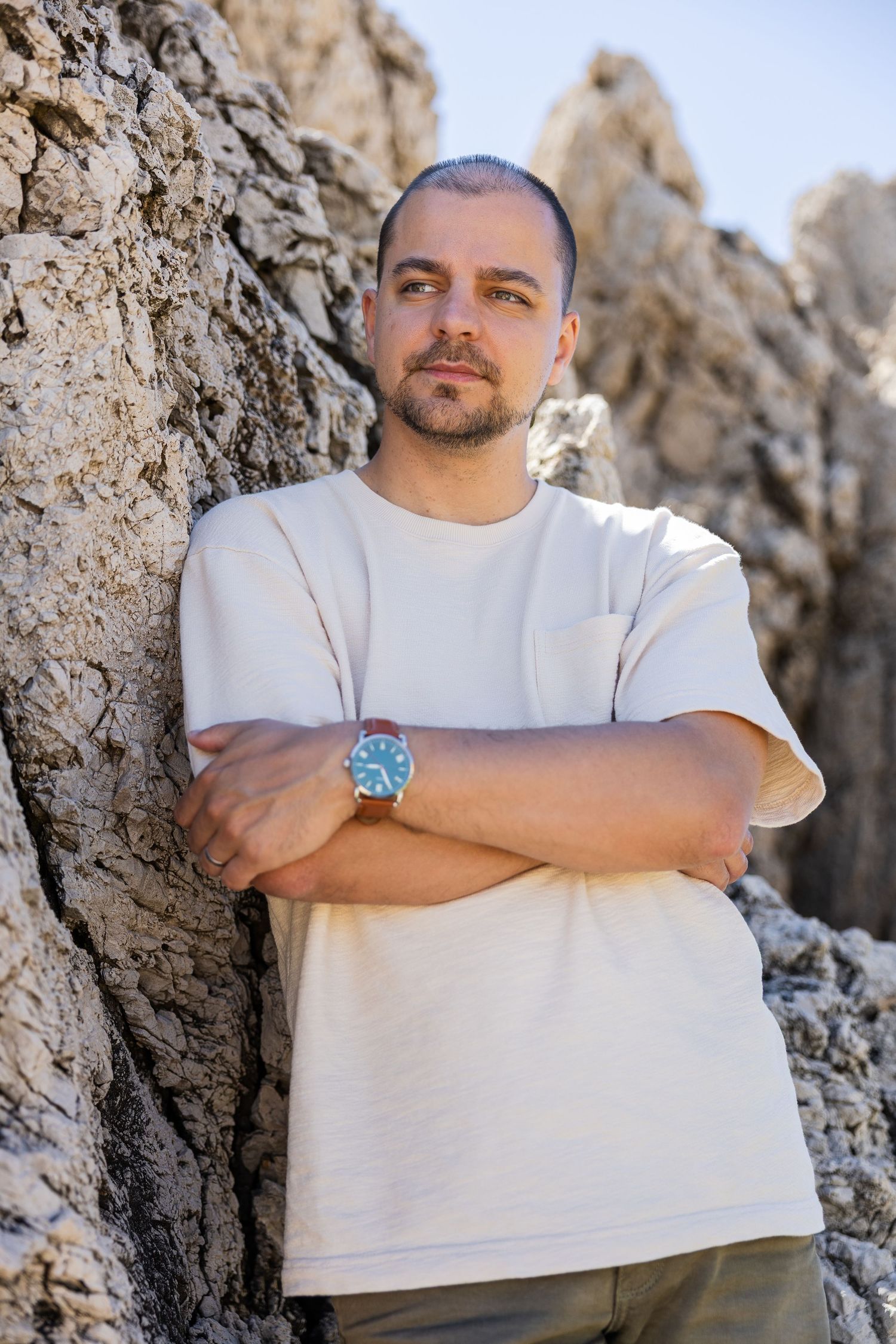 Man with crossed arms leaning against rocky cliff wearing white t-shirt and blue watch outdoors.