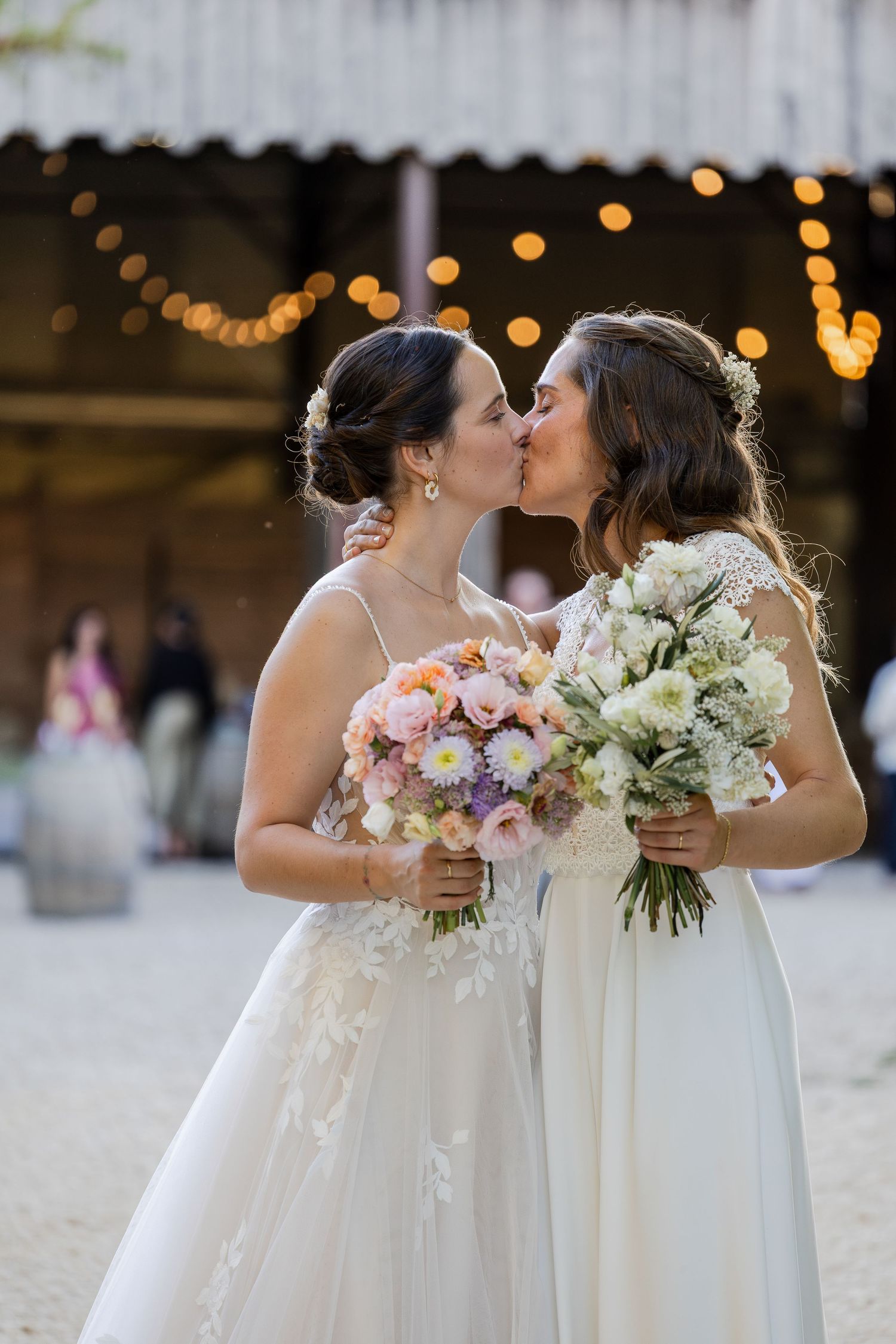 Two brides in white gowns share a kiss while holding floral bouquets at a romantic barn wedding venue.
